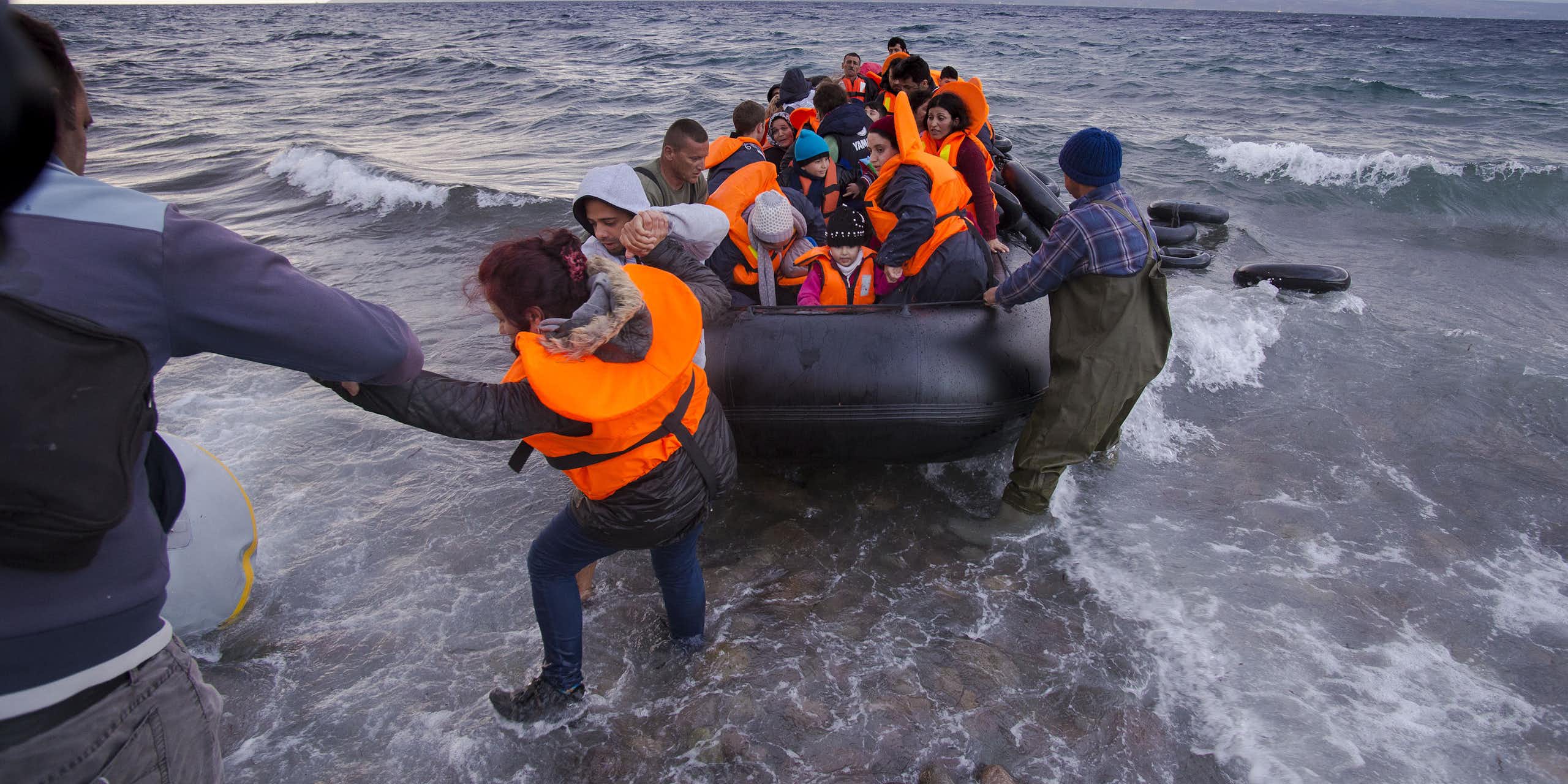 People on shore help pull a rubber dinghy full of people in life jackets from the ocean