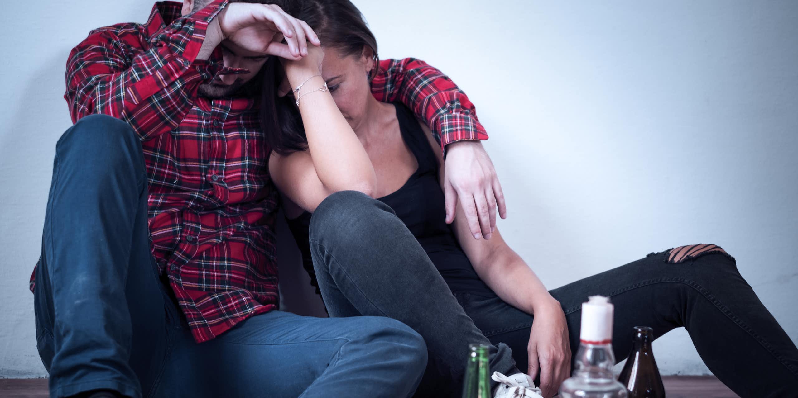 Two young people sit on the floor leaning against each other, with three empty alcohol bottles next to them.
