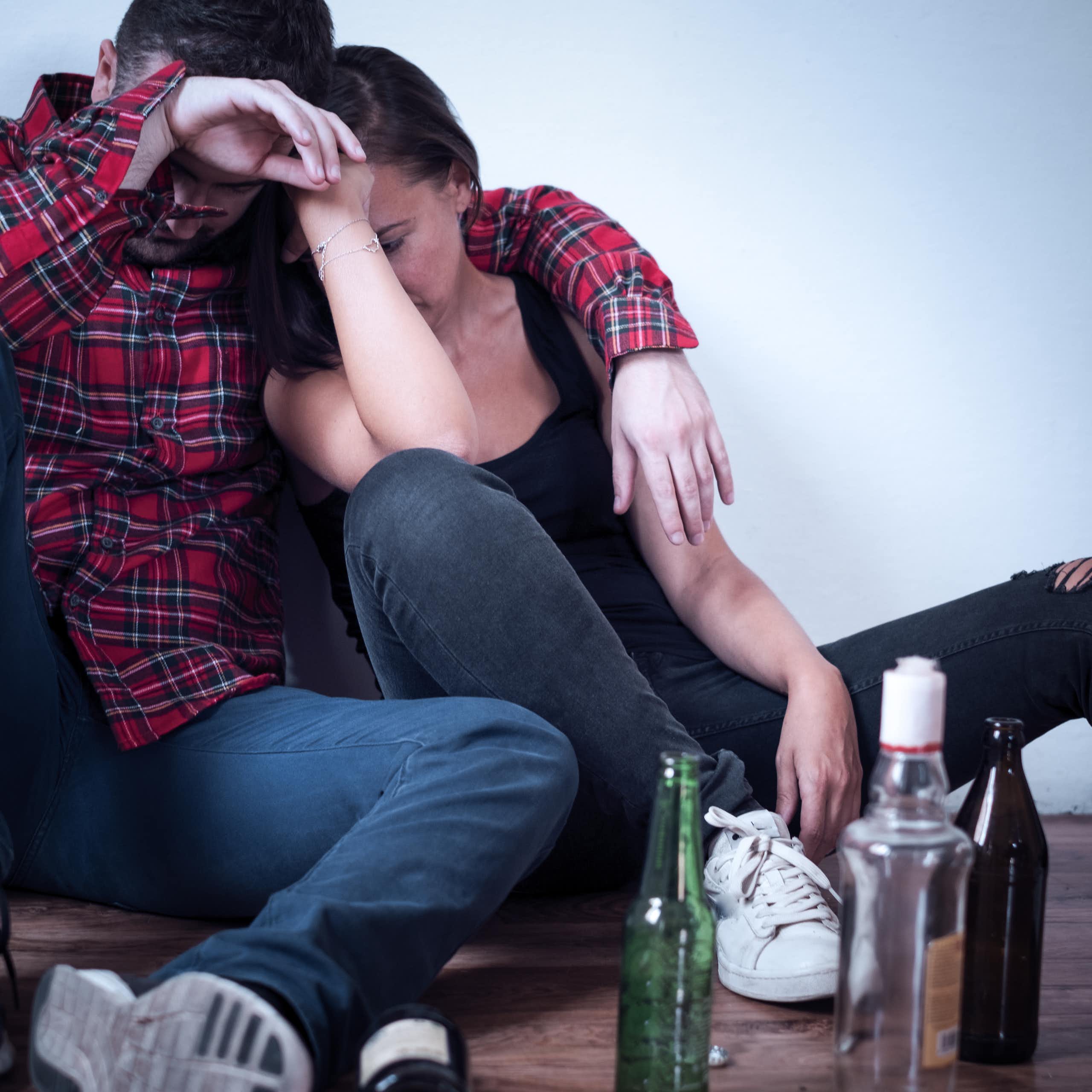 Two young people sit on the floor leaning against each other, with three empty alcohol bottles next to them.
