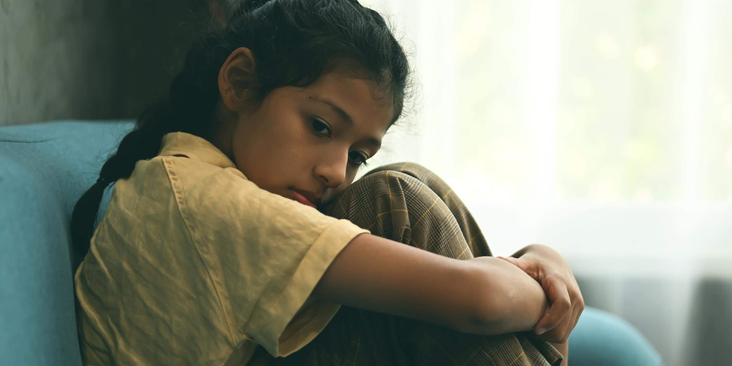 Sad girl hugs knees sitting on sofa