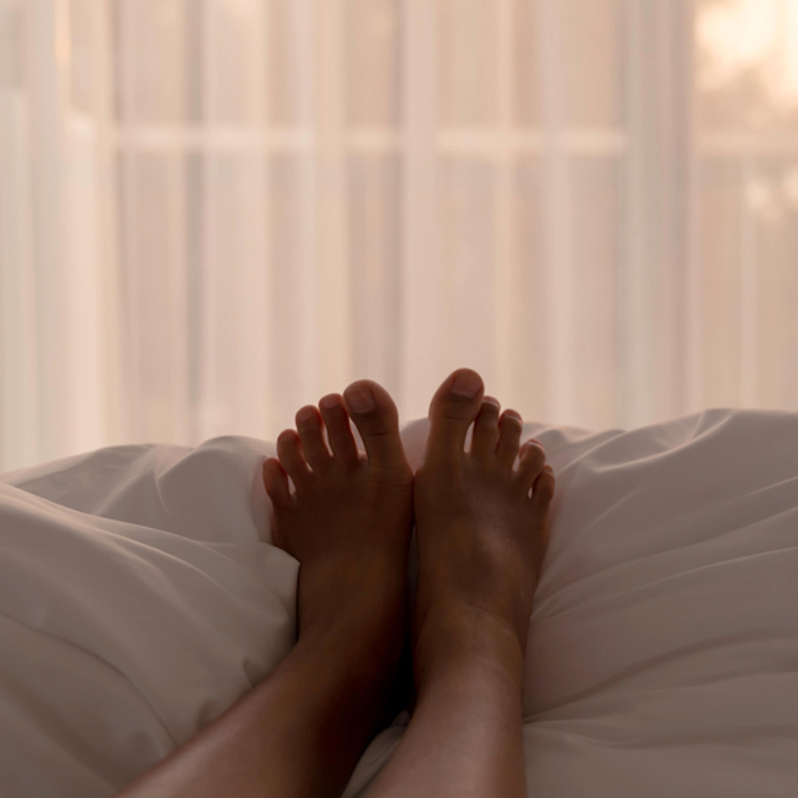 Person lying on bed, legs on top of covers, facing window, morning light shining through net curtains