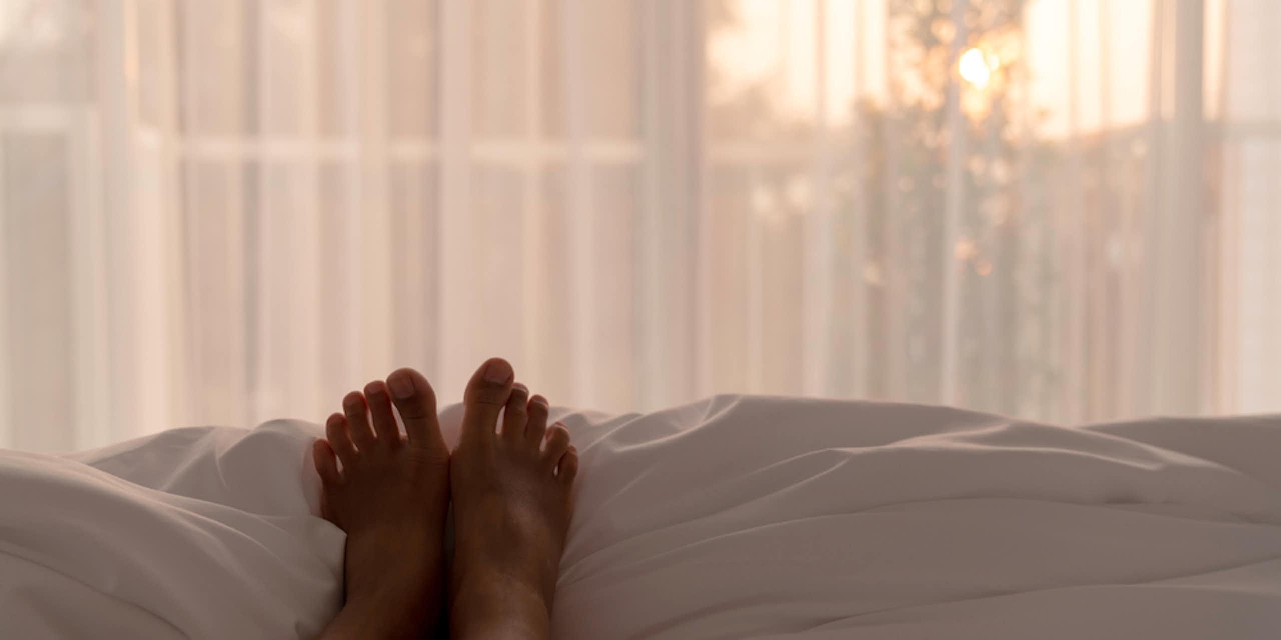 Person lying on bed, legs on top of covers, facing window, morning light shining through net curtains