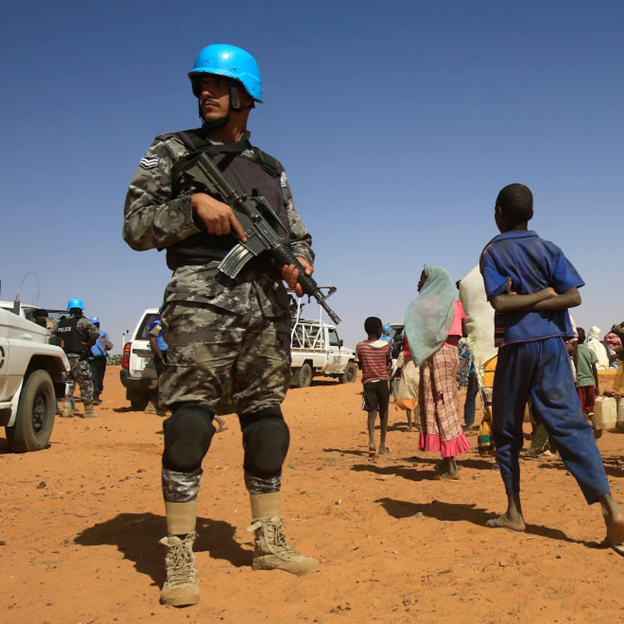 Man in camouflage uniform and blue helmet, holding a rifle, standing near a group of civilians including children
