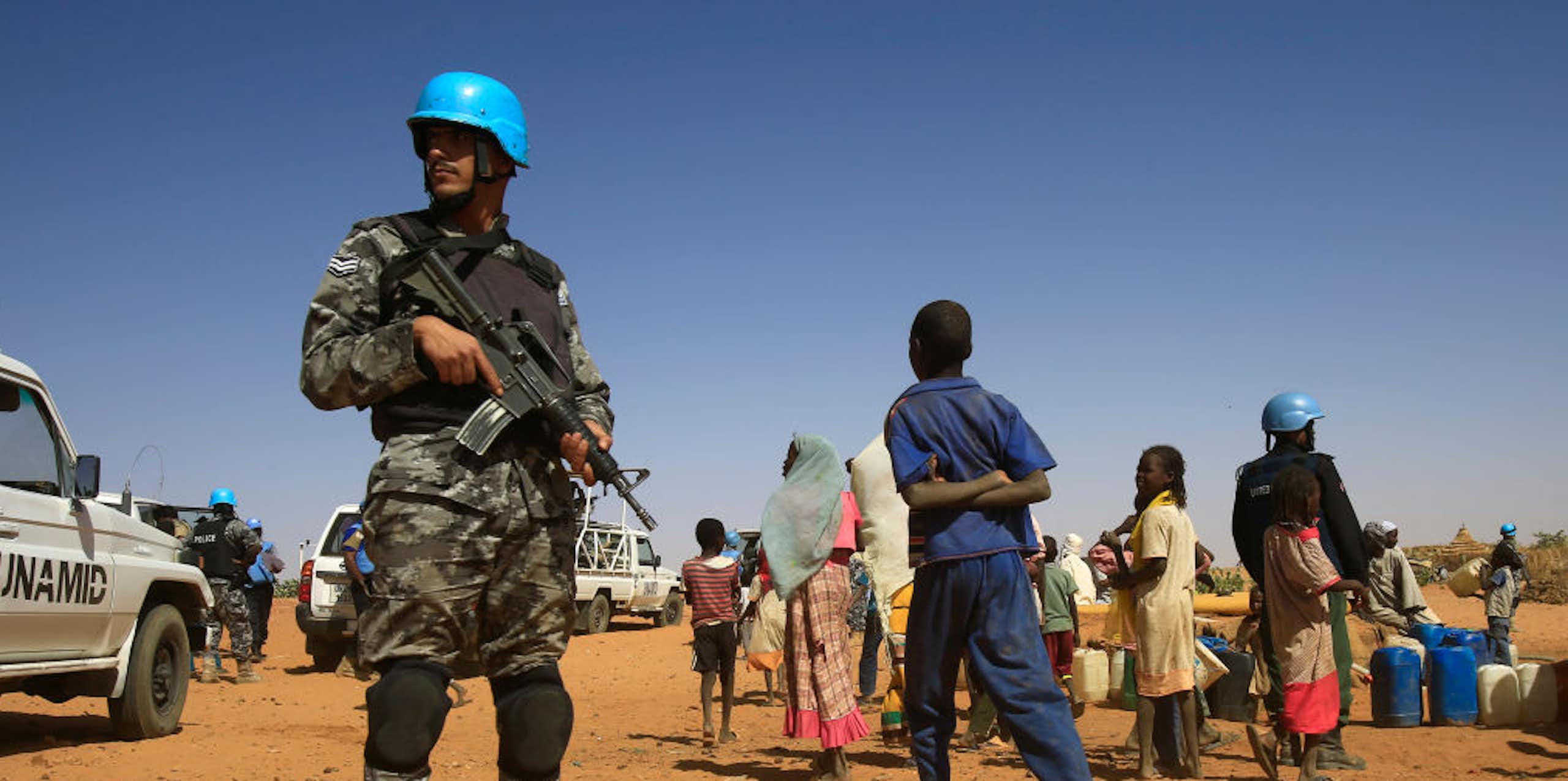 Man in camouflage uniform and blue helmet, holding a rifle, standing near a group of civilians including children