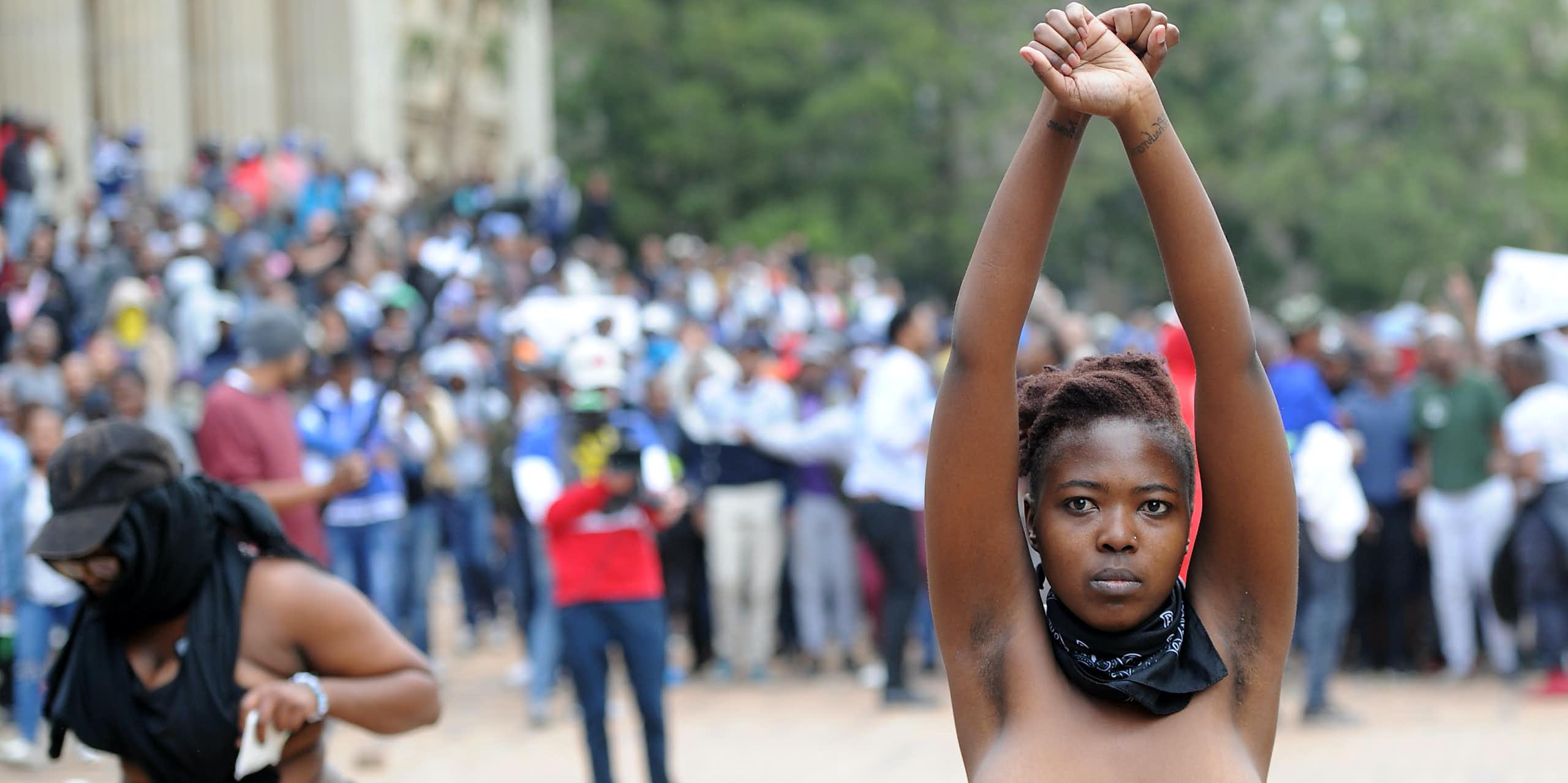 A woman stands, topless with arms raised in the air and crossed as if shackled, a bandana around her neck and a determined look on her face. Behind her, a crowd of people and a cameraman.
