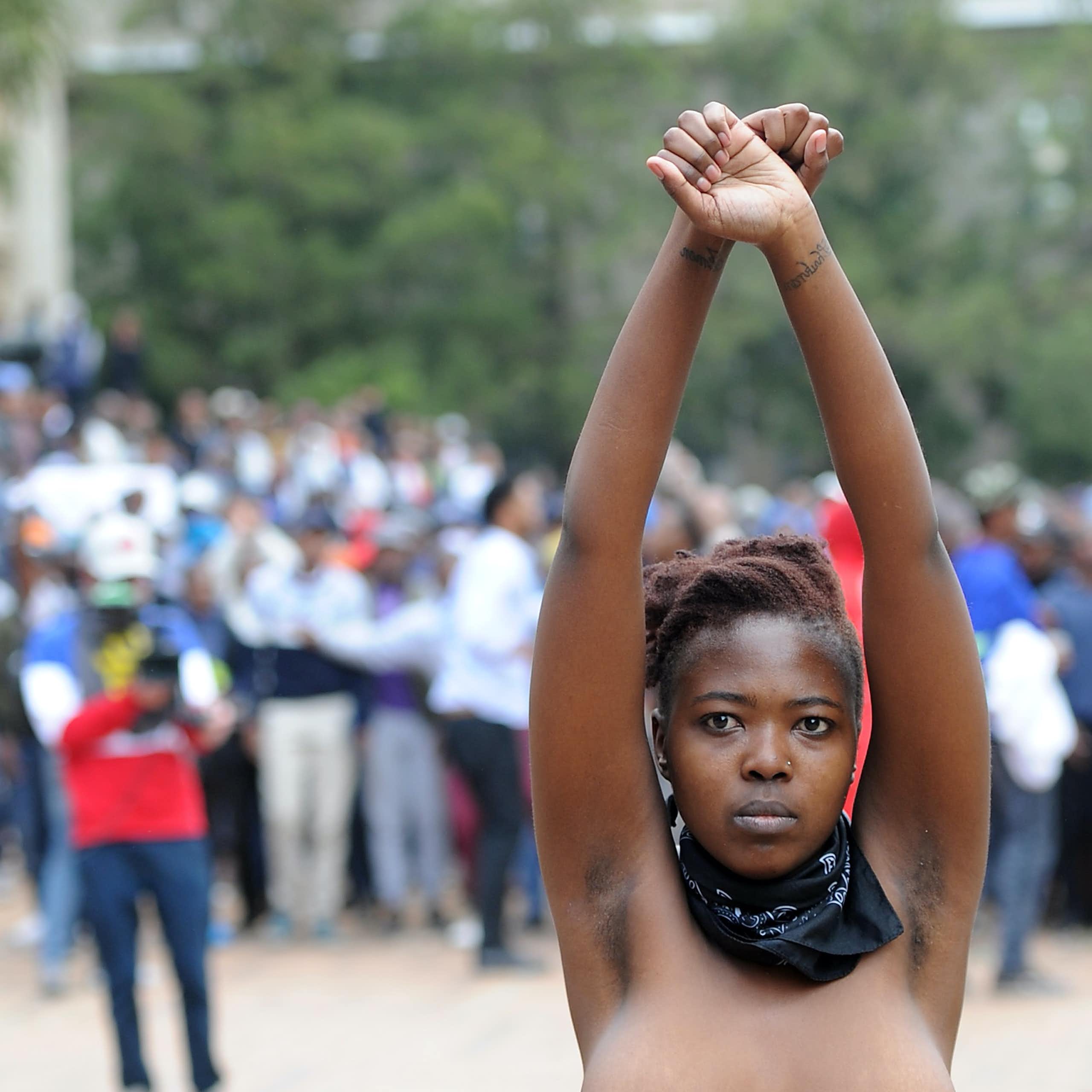 A woman stands, topless with arms raised in the air and crossed as if shackled, a bandana around her neck and a determined look on her face. Behind her, a crowd of people and a cameraman.