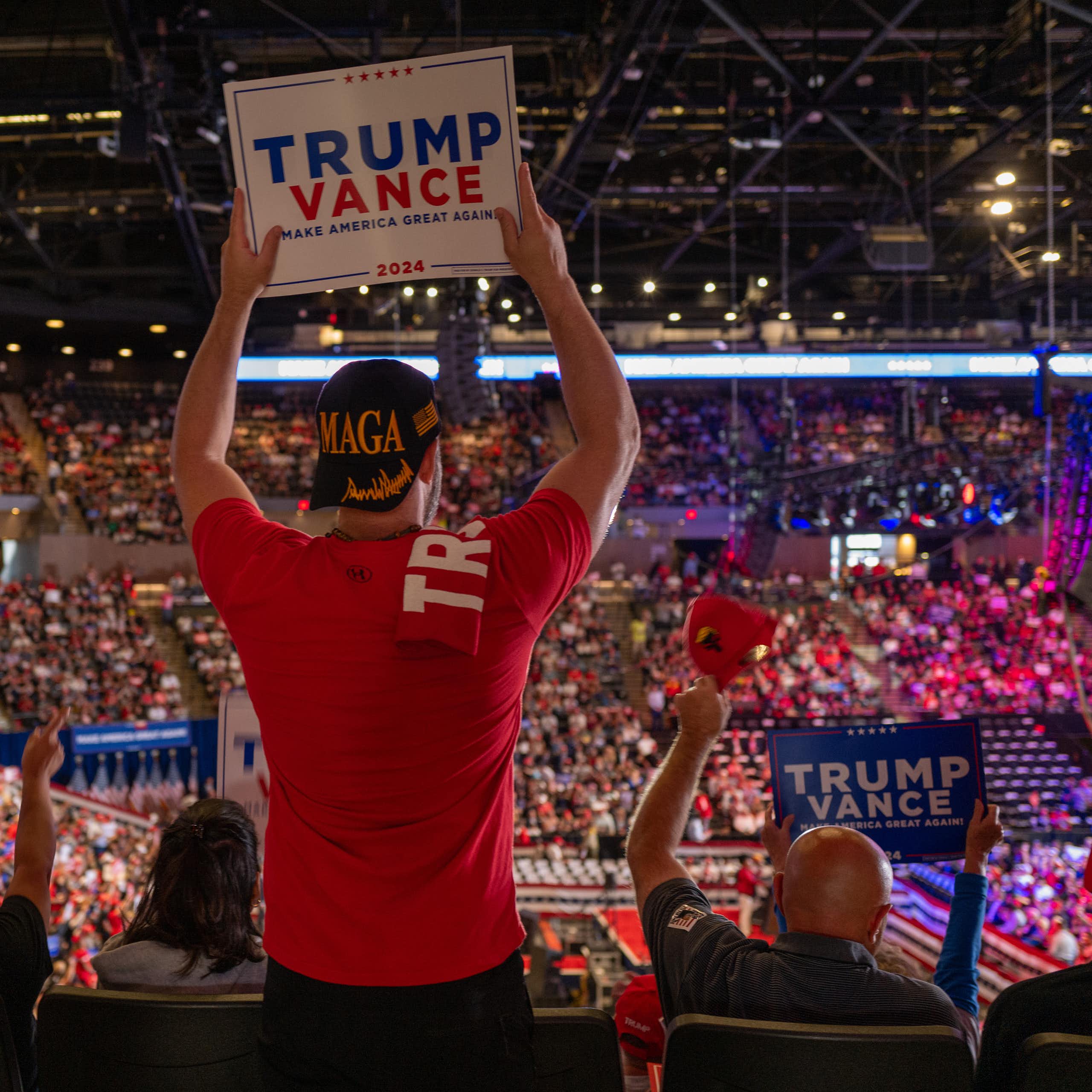 A man looks to a large crowd and wears a red shirt and holds a sign that says 'Trump Vance.' The stadium is filled with people and others hold signs that say 'Trump Vance.'