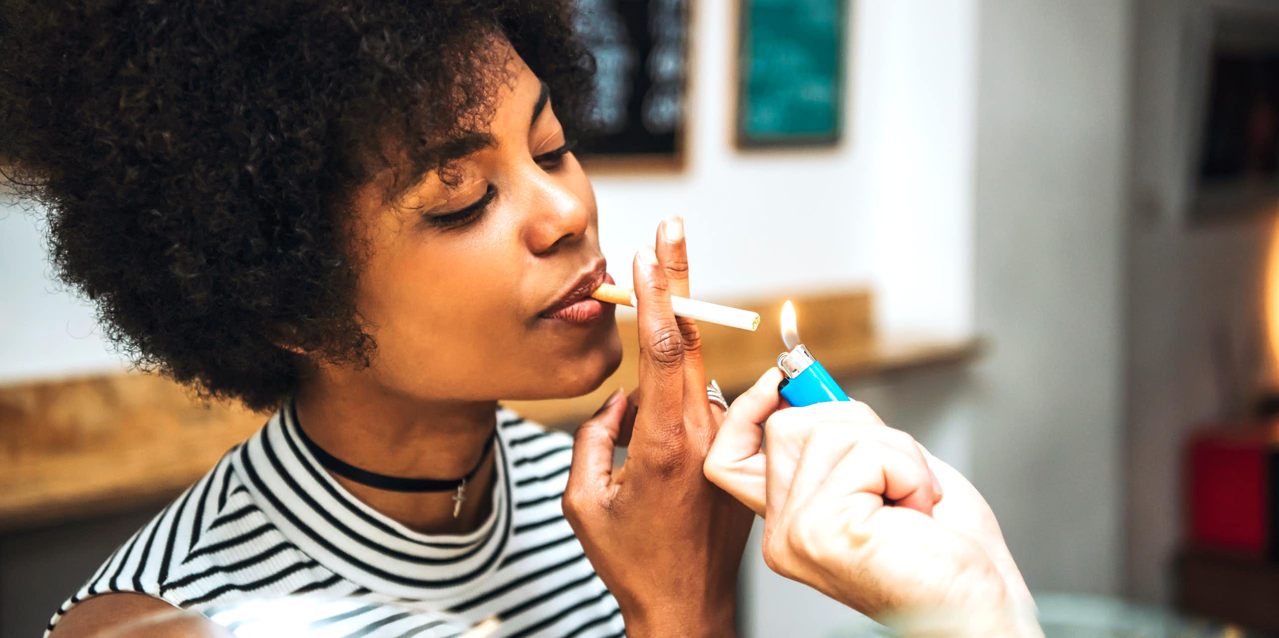 A woman with a cigarette in her mouth, accepting a light from another person
