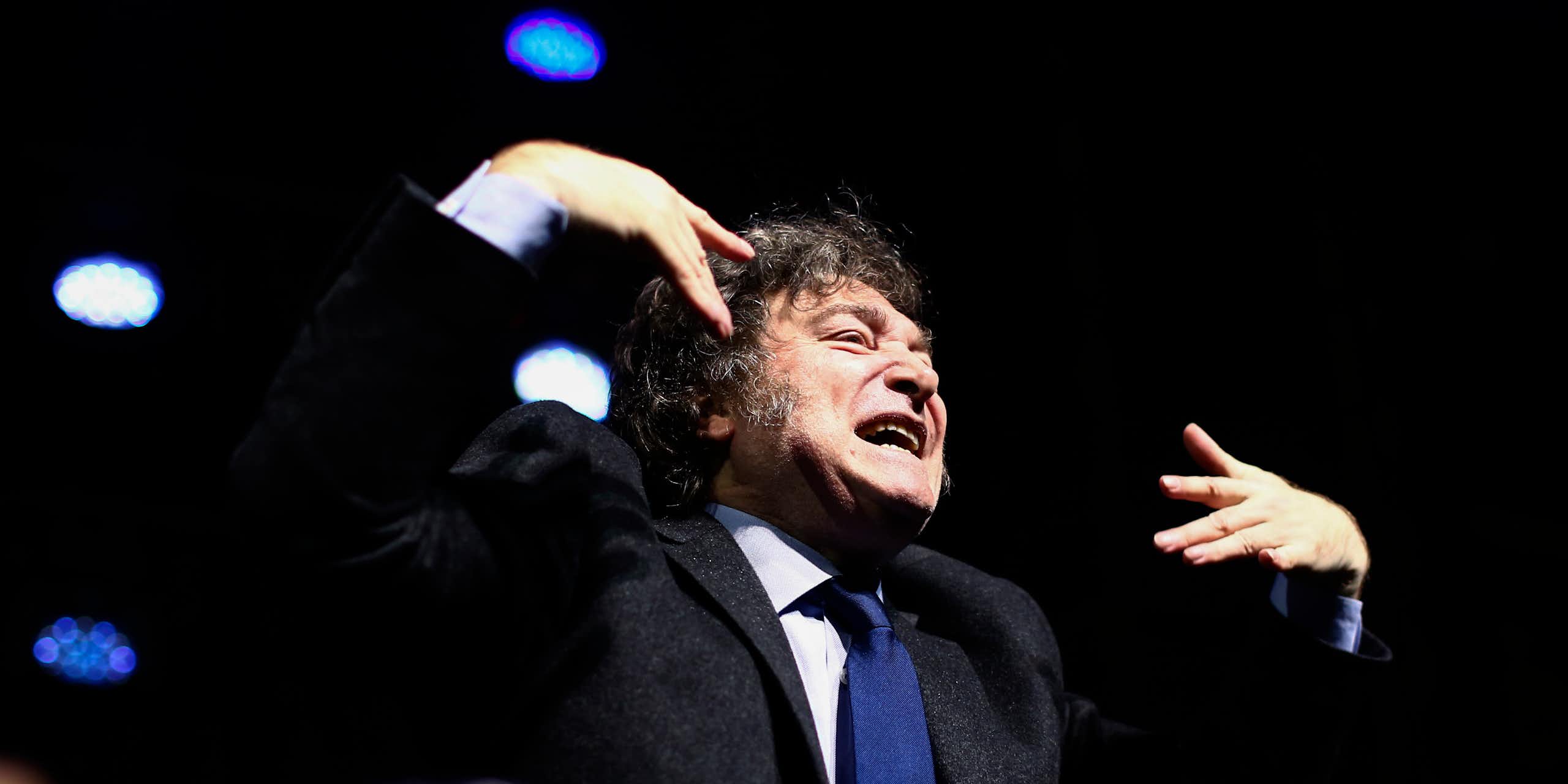 Un homme aux cheveux bruns hirsutes grimace en faisant des gestes avec ses mains.
