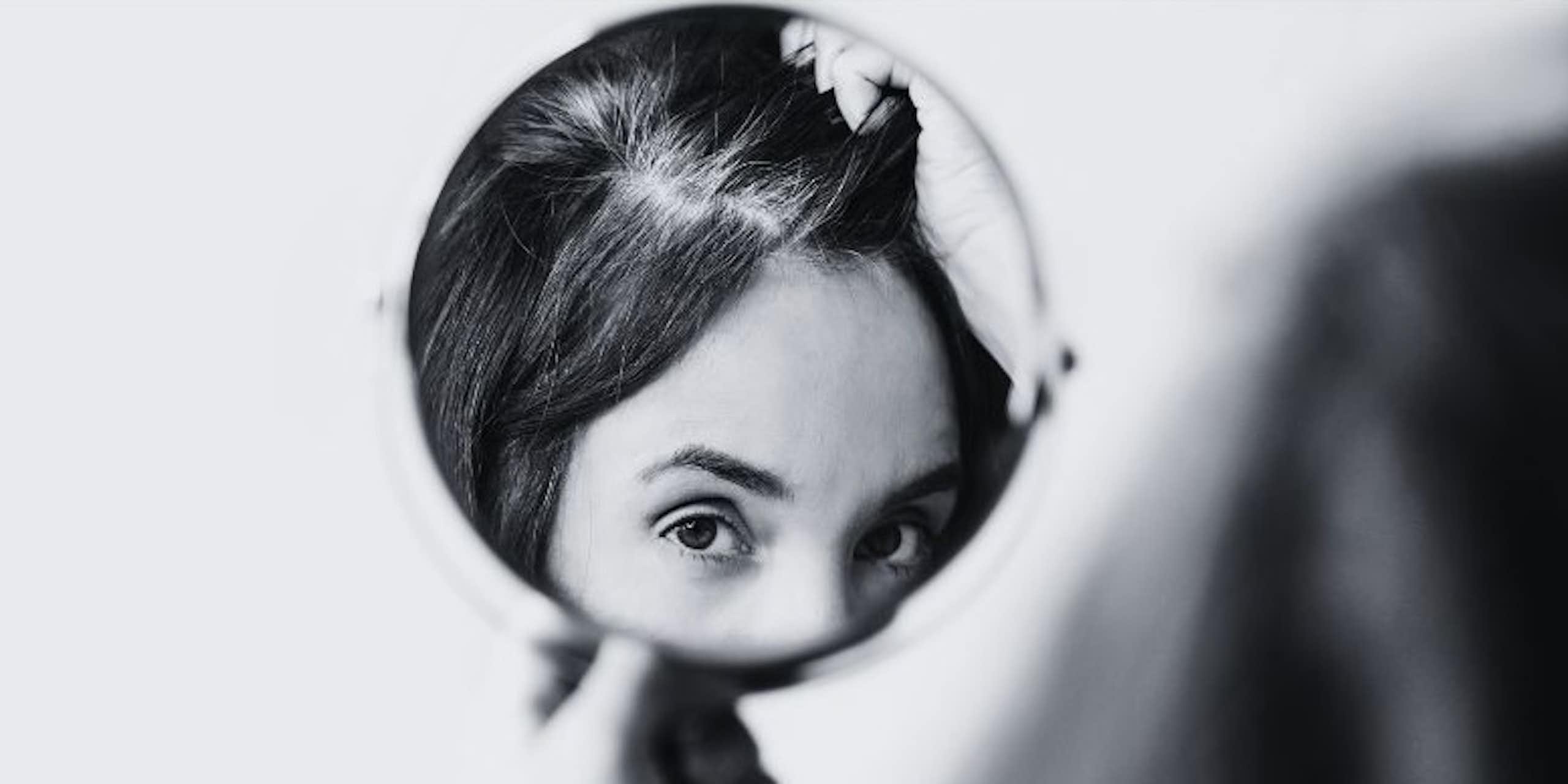 A woman inspects grey hairs in a circular mirror.