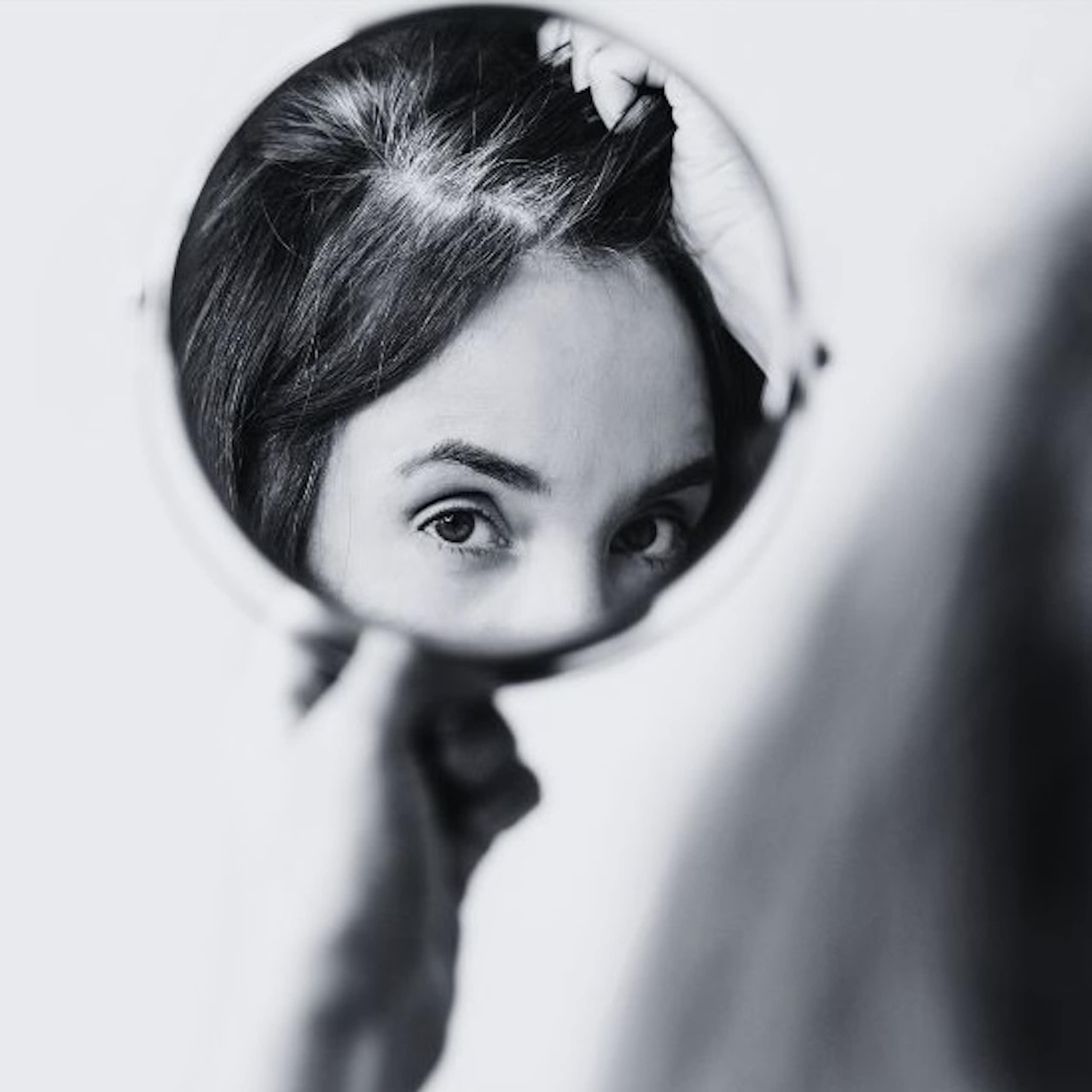 A woman inspects grey hairs in a circular mirror.