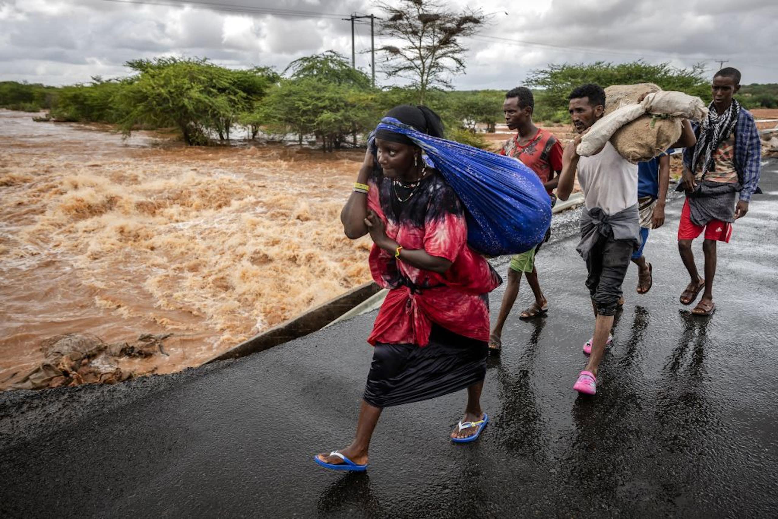 People on foot carry bundles on a wet bridge over a torrential river