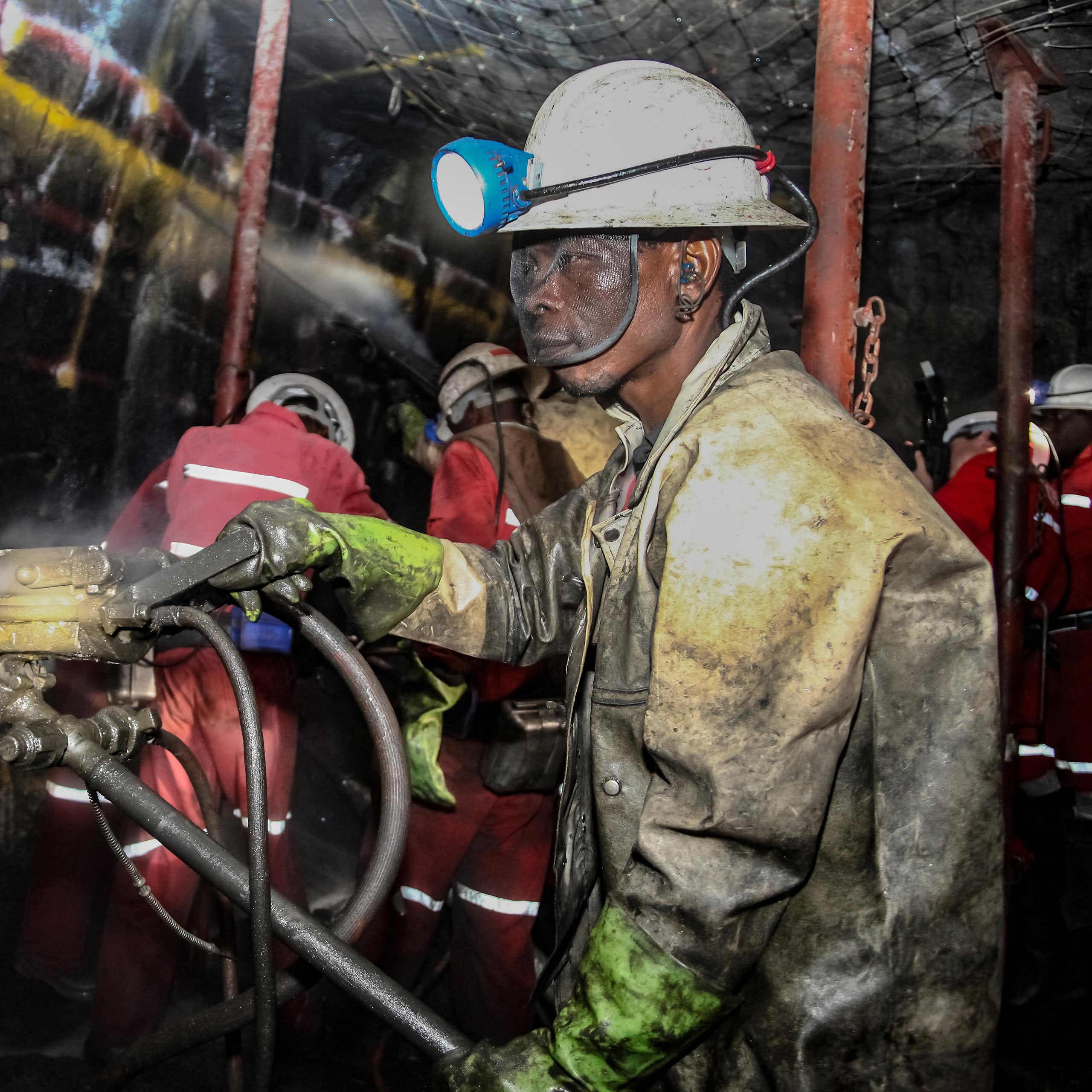 A man in a hard hat and a heavy uniform, his eyes protected by safety goggles, works underground in a mine
