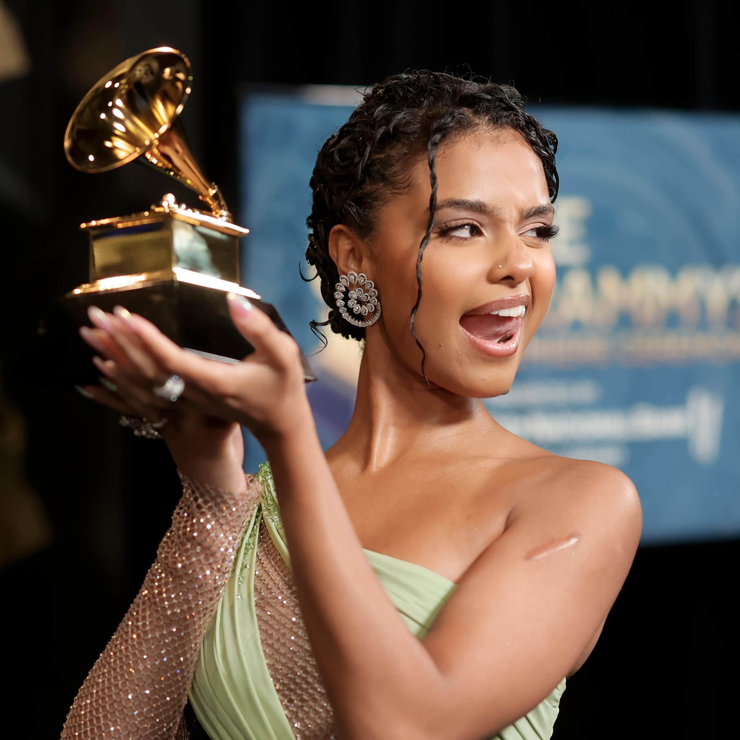 A young woman in an off-the-shoulder evening gown sticks her tongue out cheekily while displaying a large gold statue shaped like a gramophone