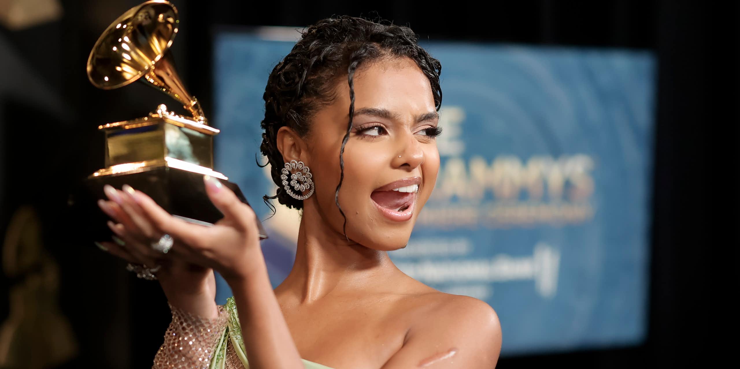 A young woman in an off-the-shoulder evening gown sticks her tongue out cheekily while displaying a large gold statue shaped like a gramophone
