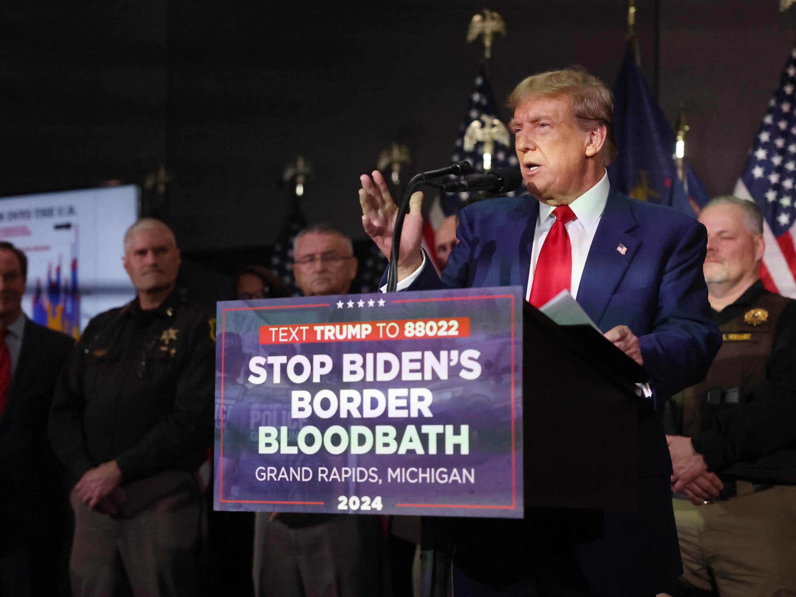 A man in a blue blazer standing at a lectern with a sign that says 'STOP BIDEN'S BORDER BLOODBATH'
