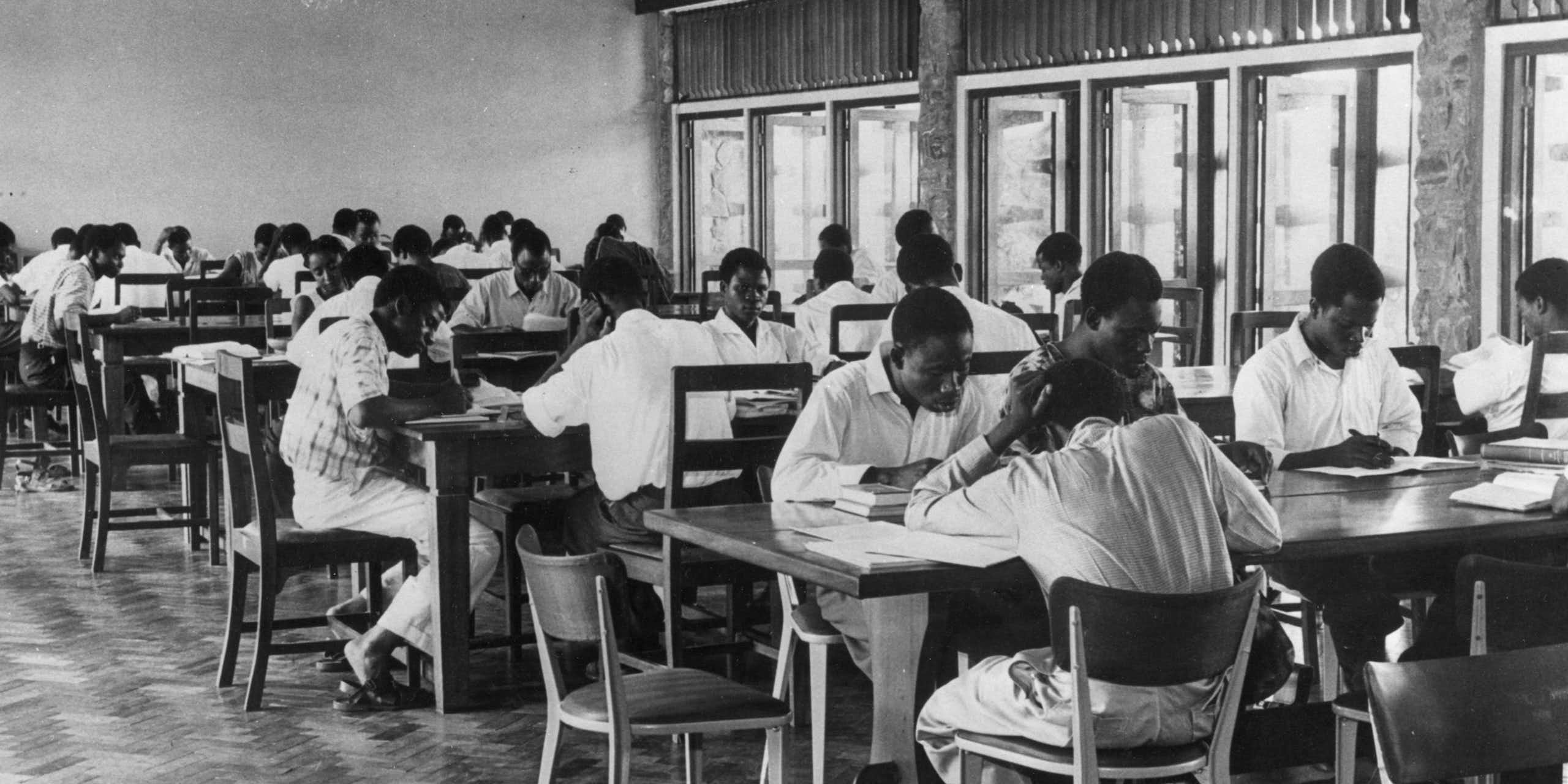 A black and white photograph of people sitting at long tables, reading