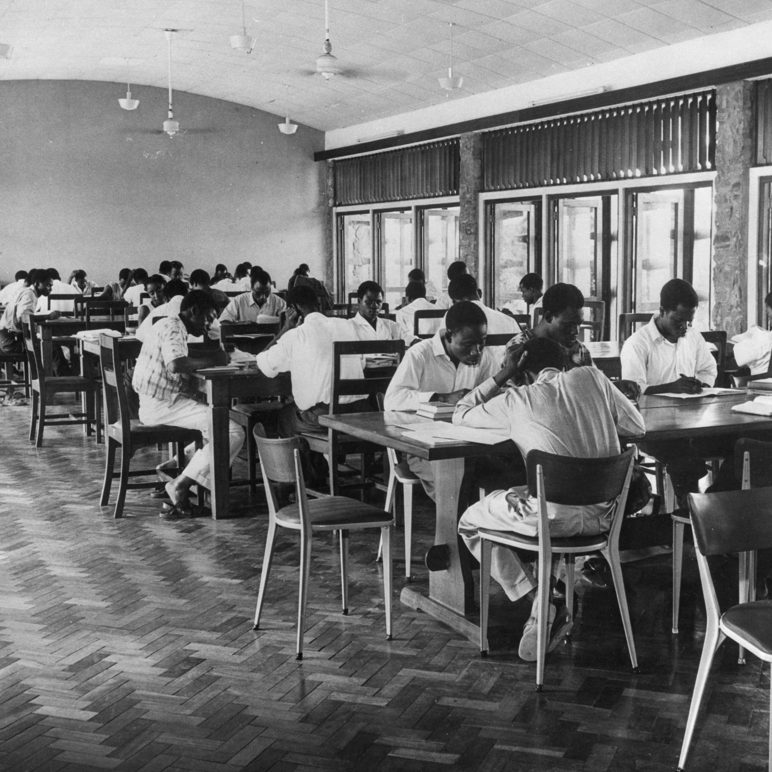 A black and white photograph of people sitting at long tables, reading