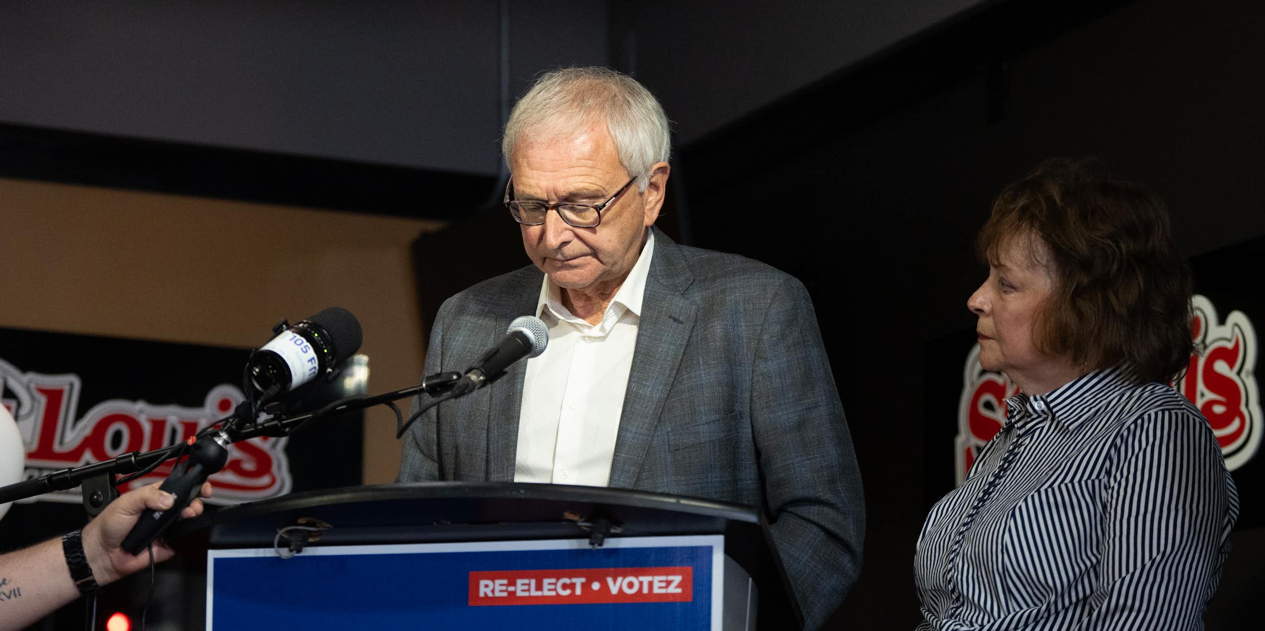 A grey-haired man in glasses looks down as he stands behind a podium that reads Re-elect Blaine Higgs. A dark-haired woman in a striped dress stands beside him.