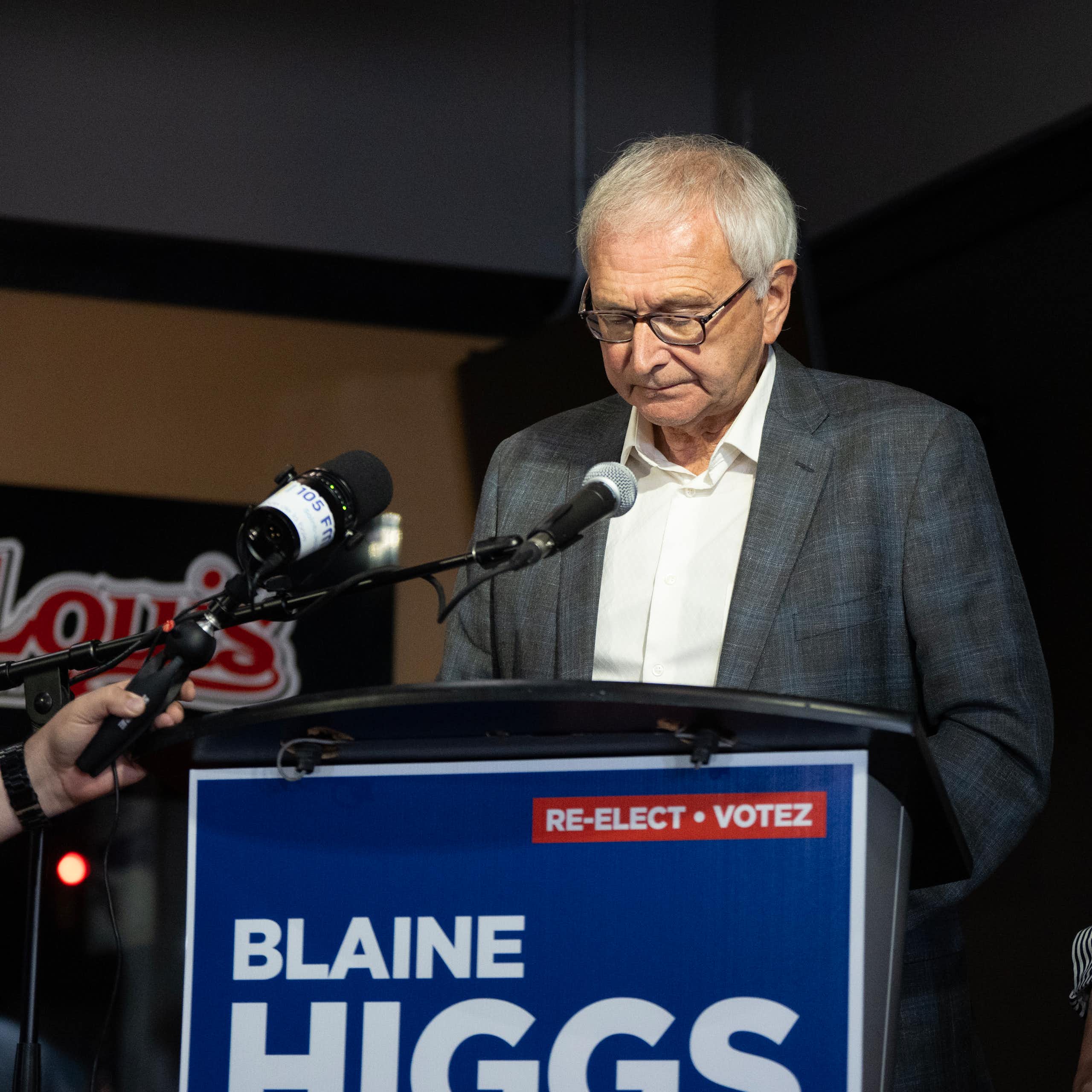 A grey-haired man in glasses looks down as he stands behind a podium that reads Re-elect Blaine Higgs. A dark-haired woman in a striped dress stands beside him.
