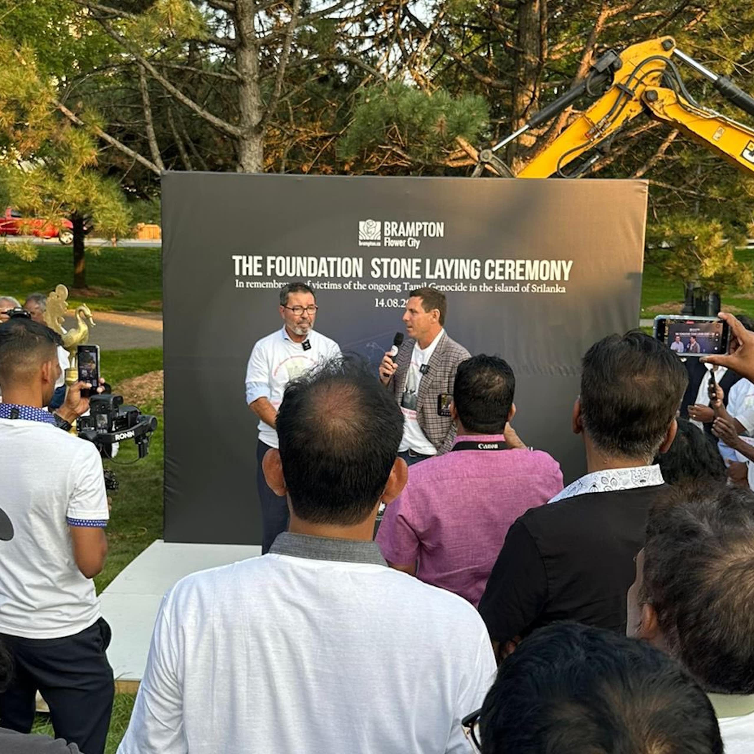 A man speaking into a mic in front of a sign reading foundation stone laying ceremony. Others are gathered around. They are standing in a park