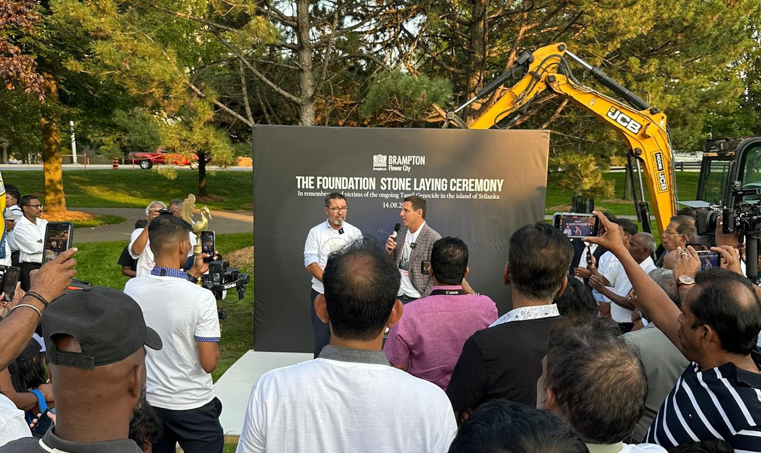 A man speaking into a mic in front of a sign reading foundation stone laying ceremony. Others are gathered around. They are standing in a park