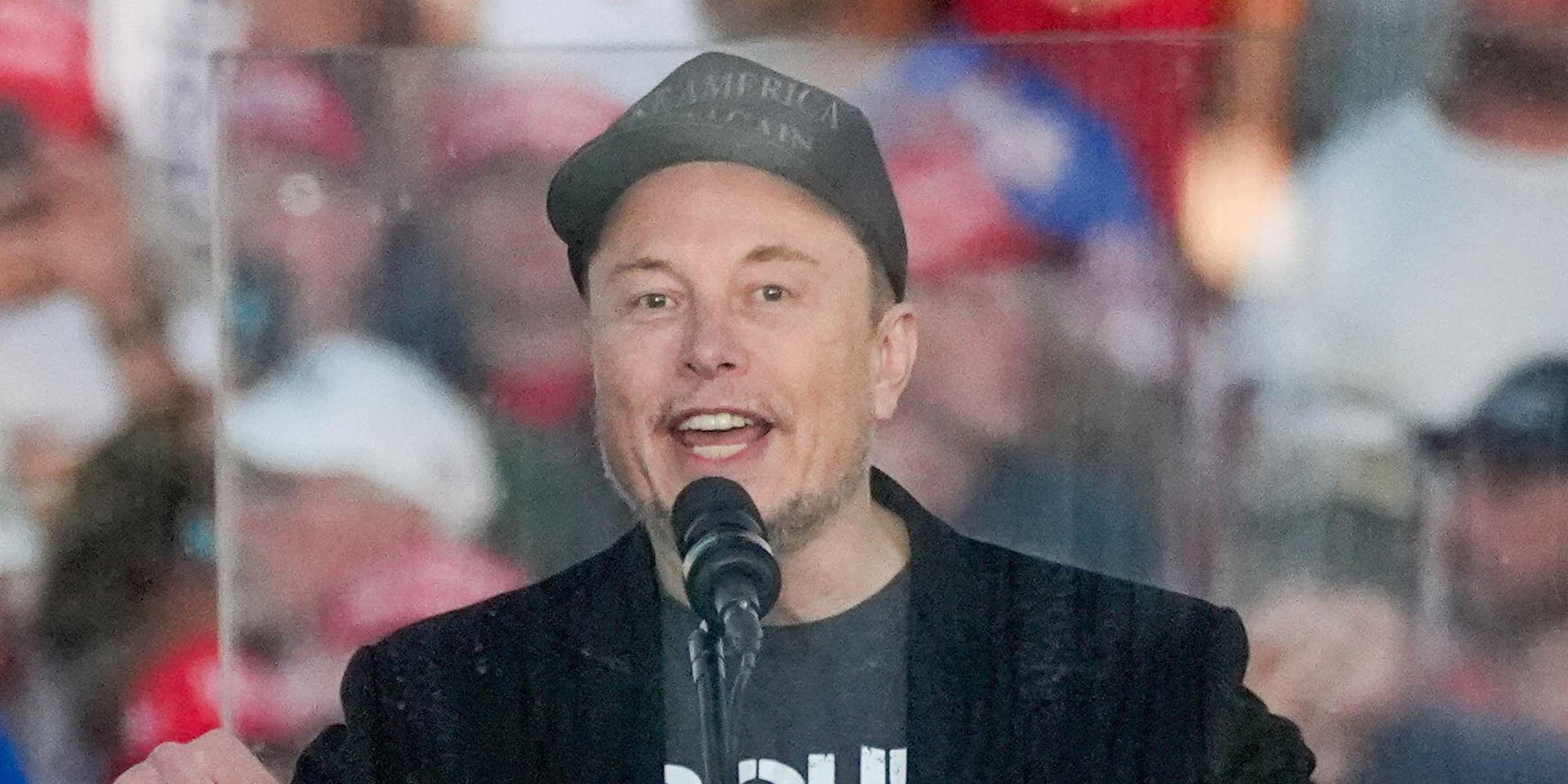 Elon Musk stands in front of a monitor speaking at a Trump rally.