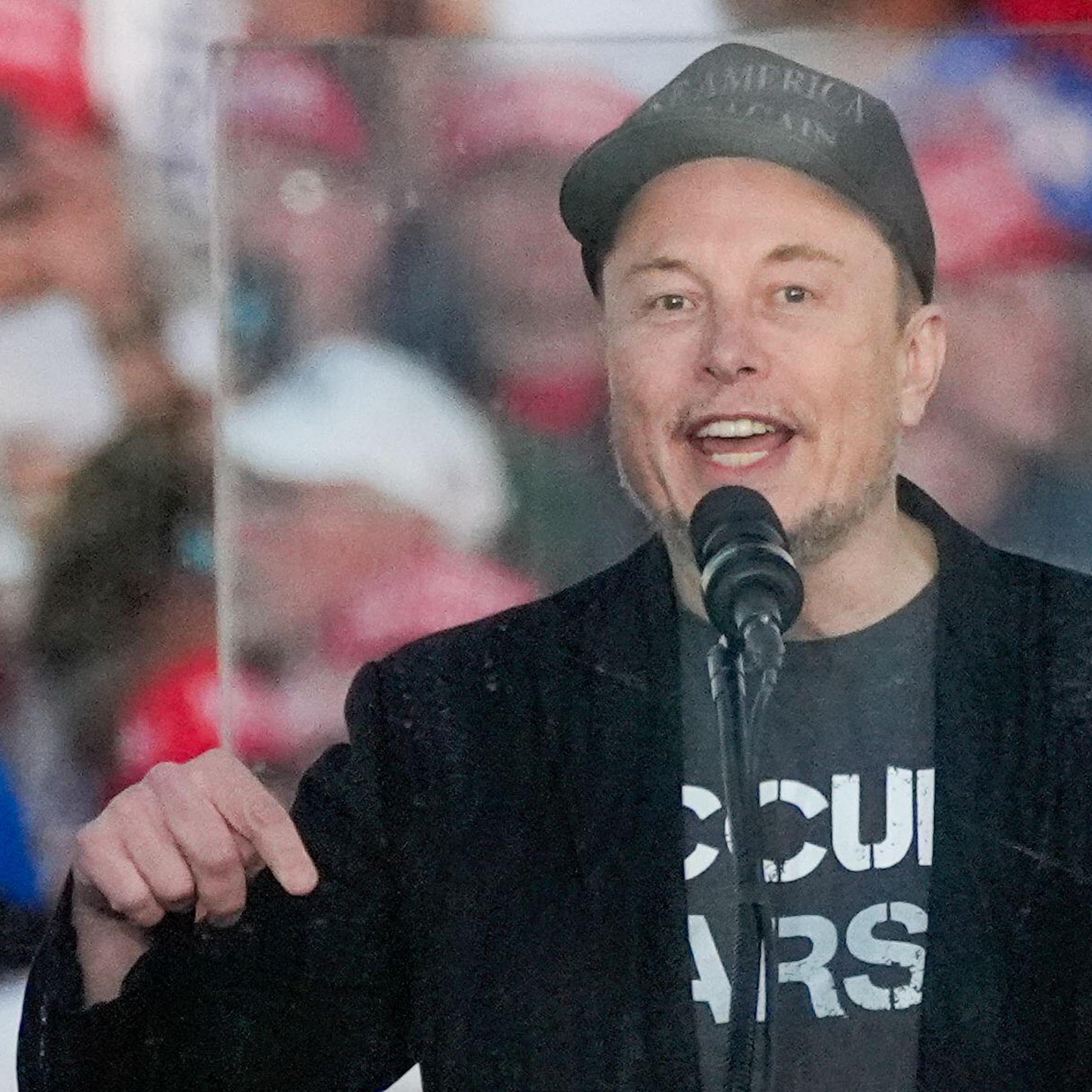 Elon Musk stands in front of a monitor speaking at a Trump rally.
