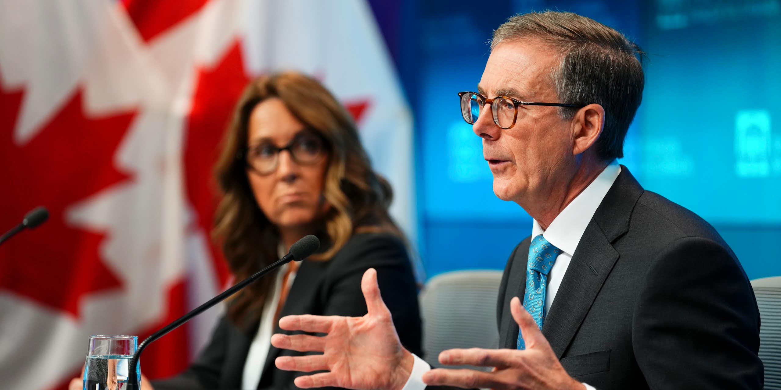 An older white man in glasses gestures with his hands while speaking and looking off-camera. Beside him sits a white woman with wavy brown hair and glasses.