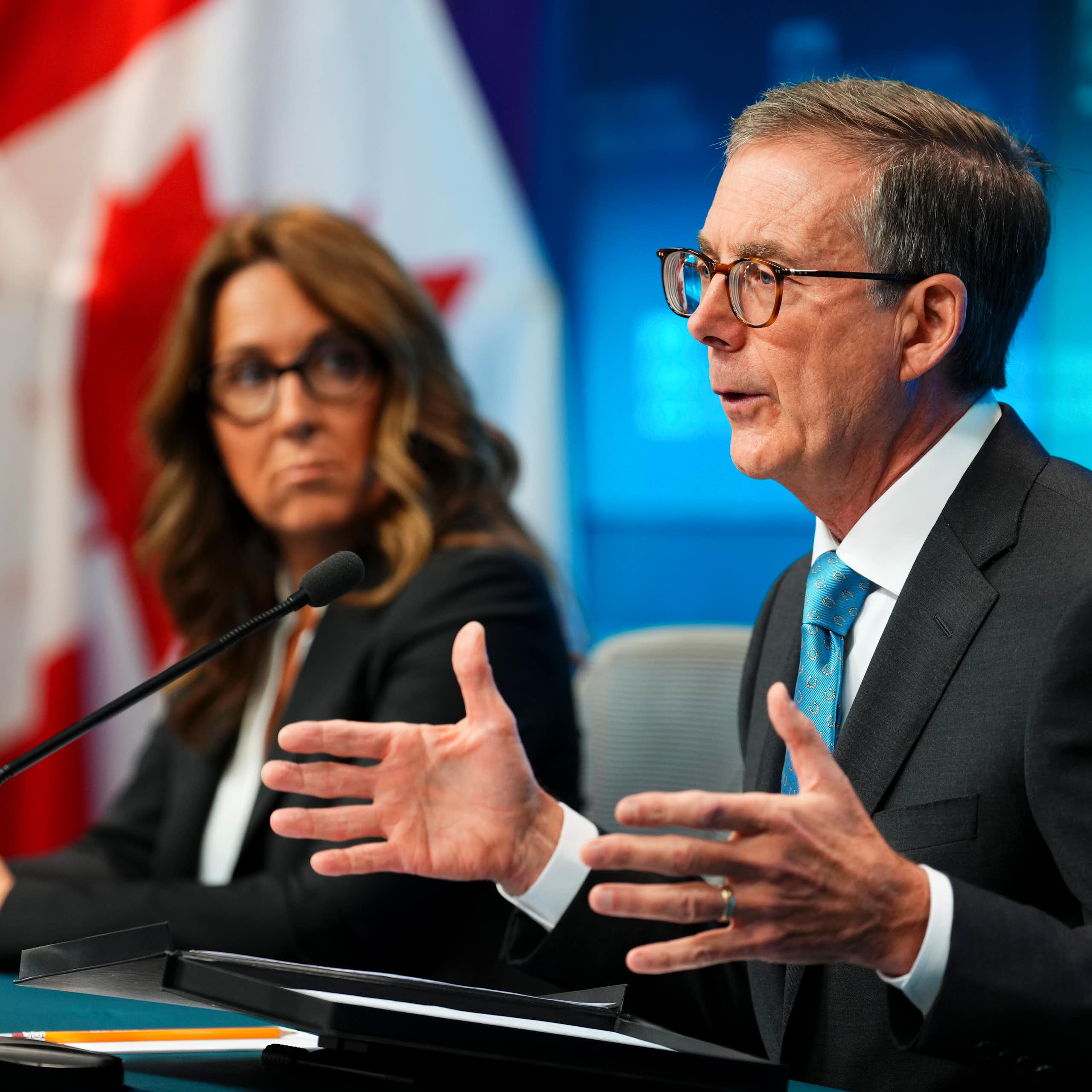 An older white man in glasses gestures with his hands while speaking and looking off-camera. Beside him sits a white woman with wavy brown hair and glasses.