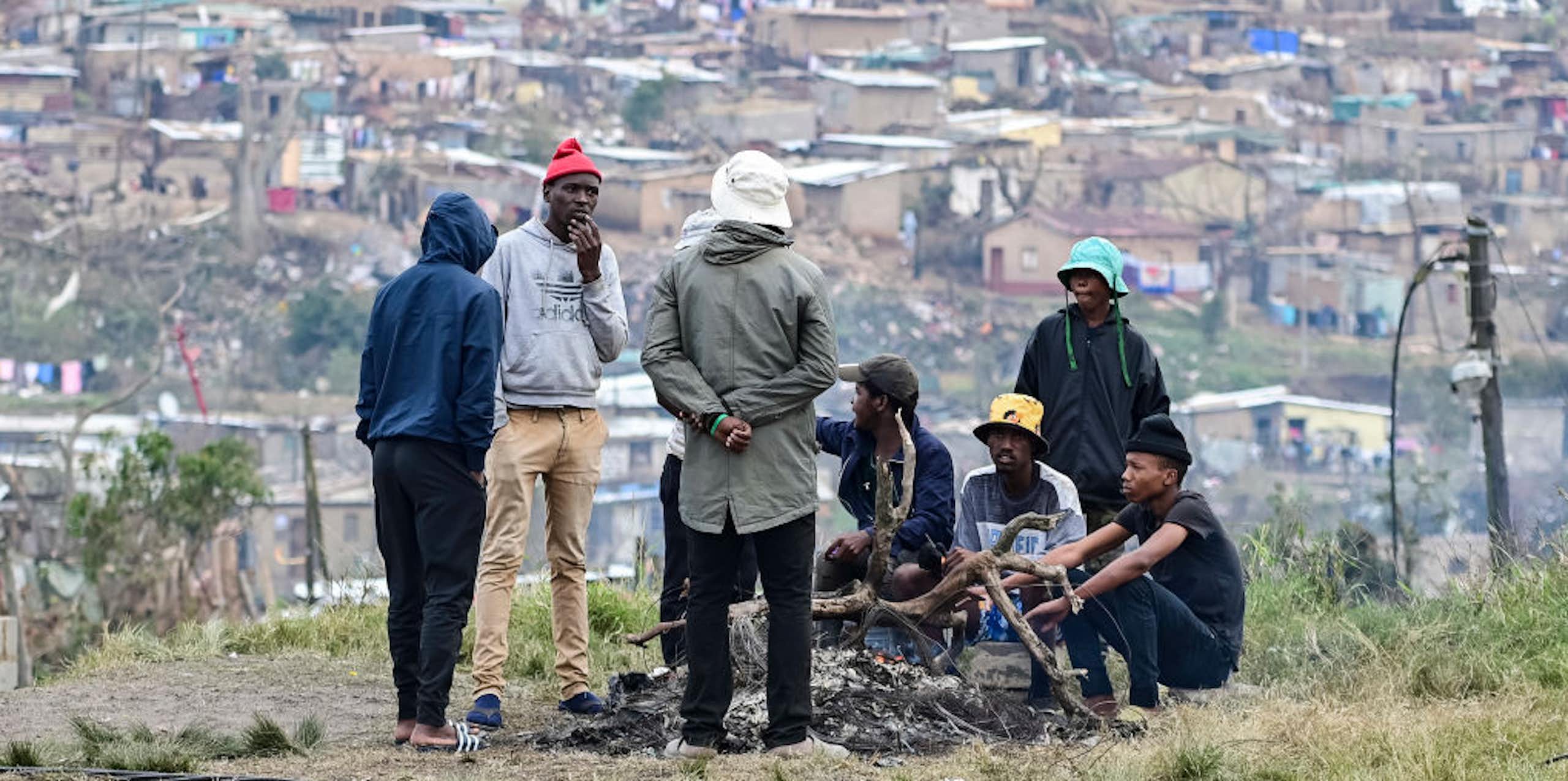 Young men around a fire on open ground. A dense settlement in the background.