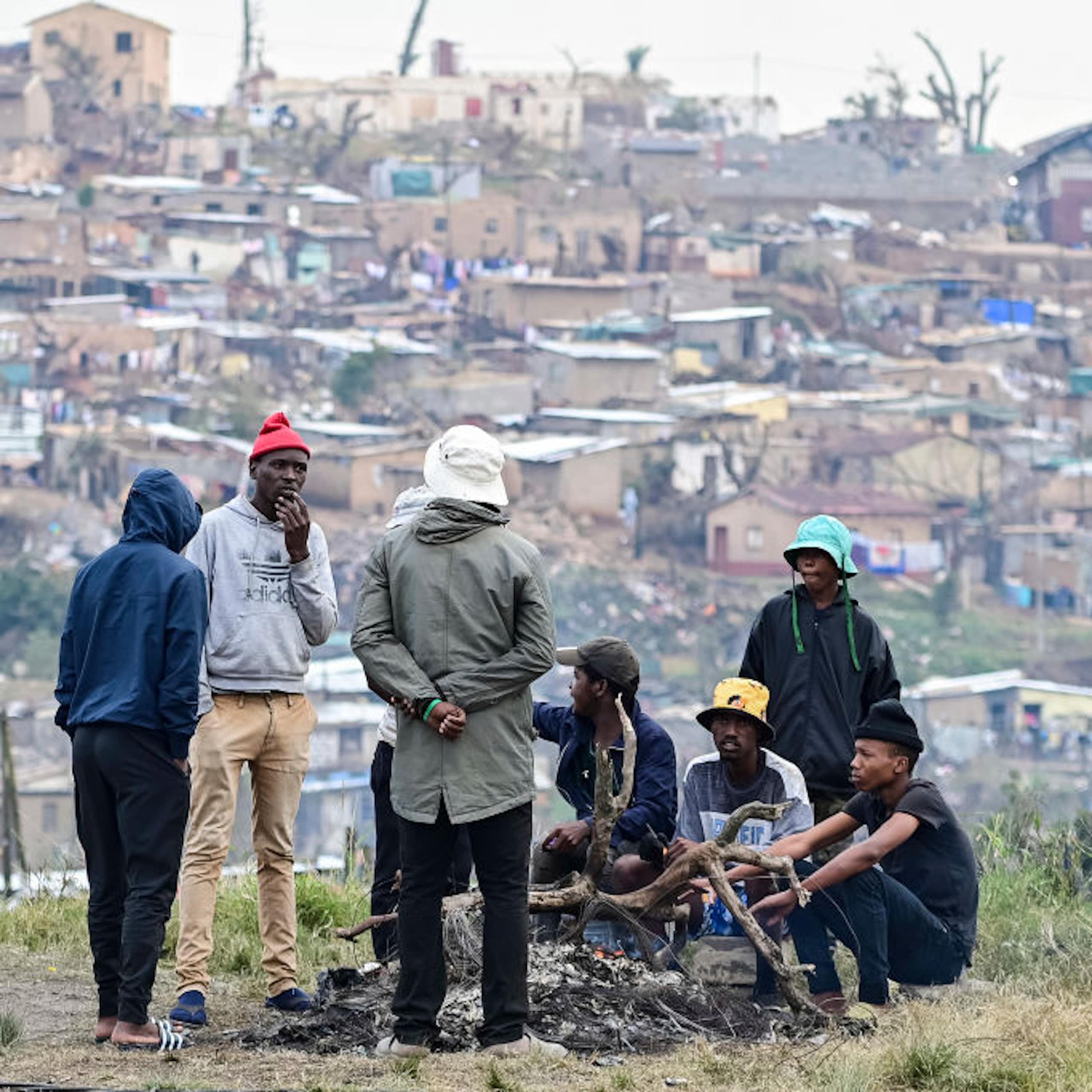 Young men around a fire on open ground. A dense settlement in the background.