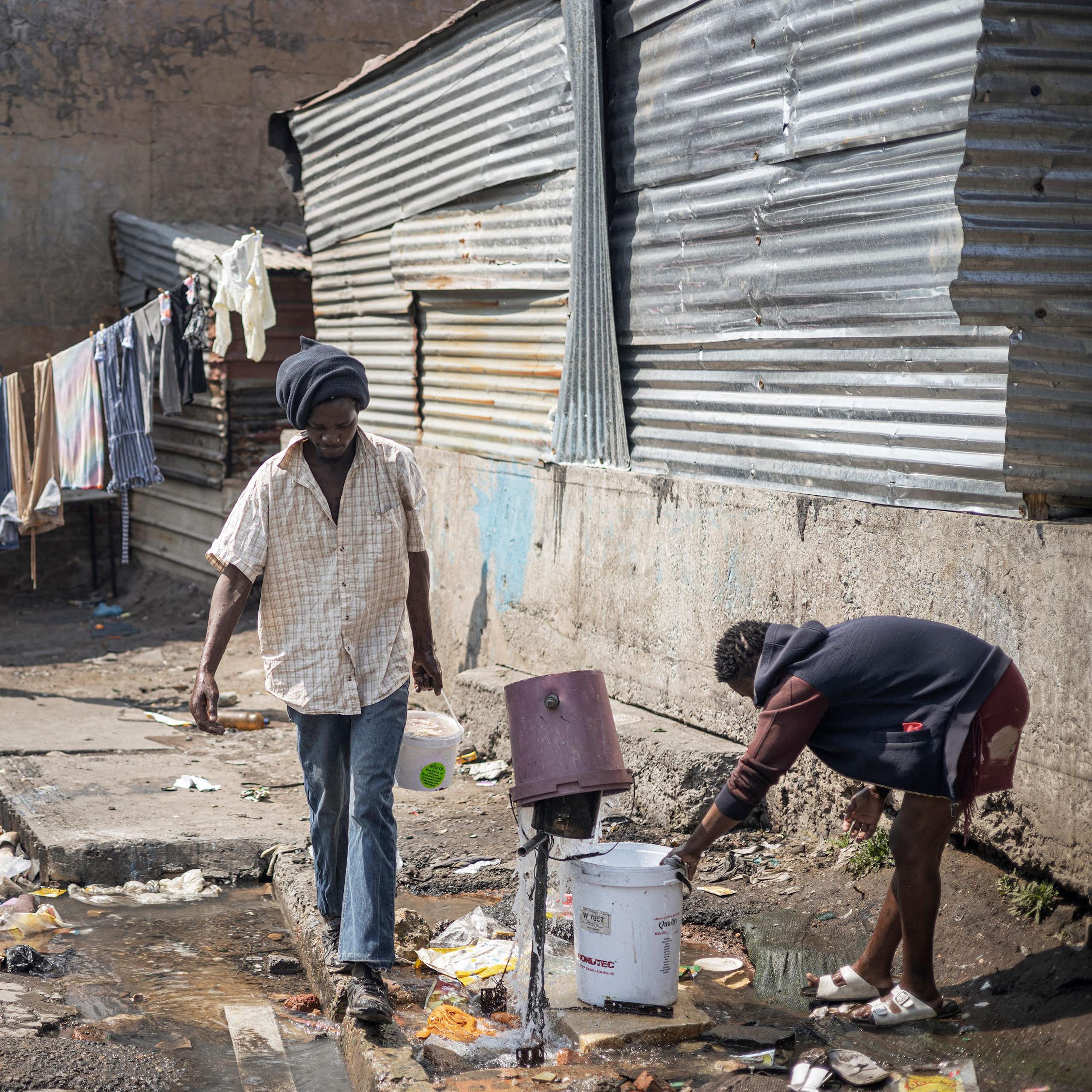 A broken tap with water spurting in different directions and a woman trying to collect the water with a bucket