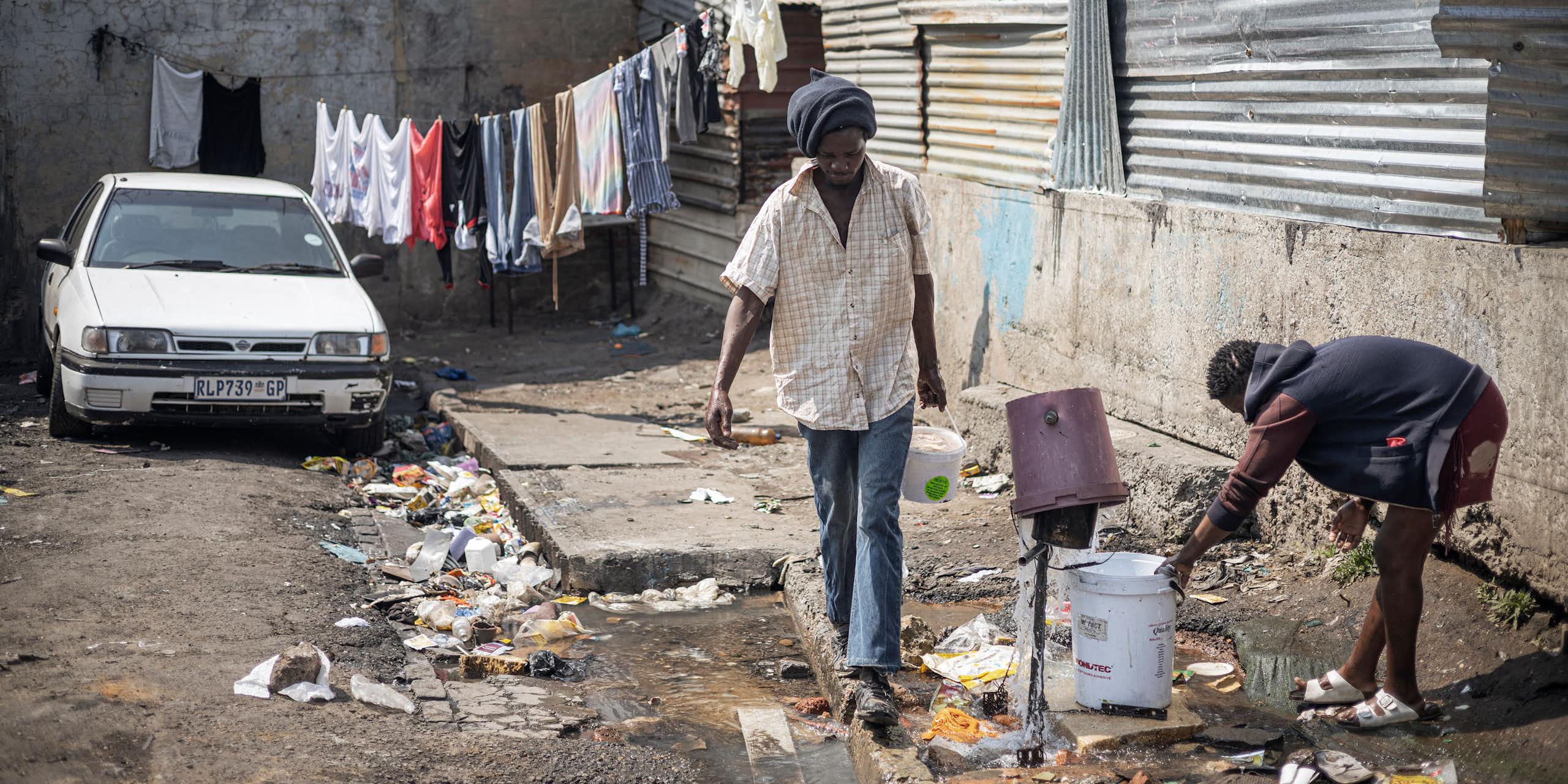 A broken tap with water spurting in different directions and a woman trying to collect the water with a bucket
