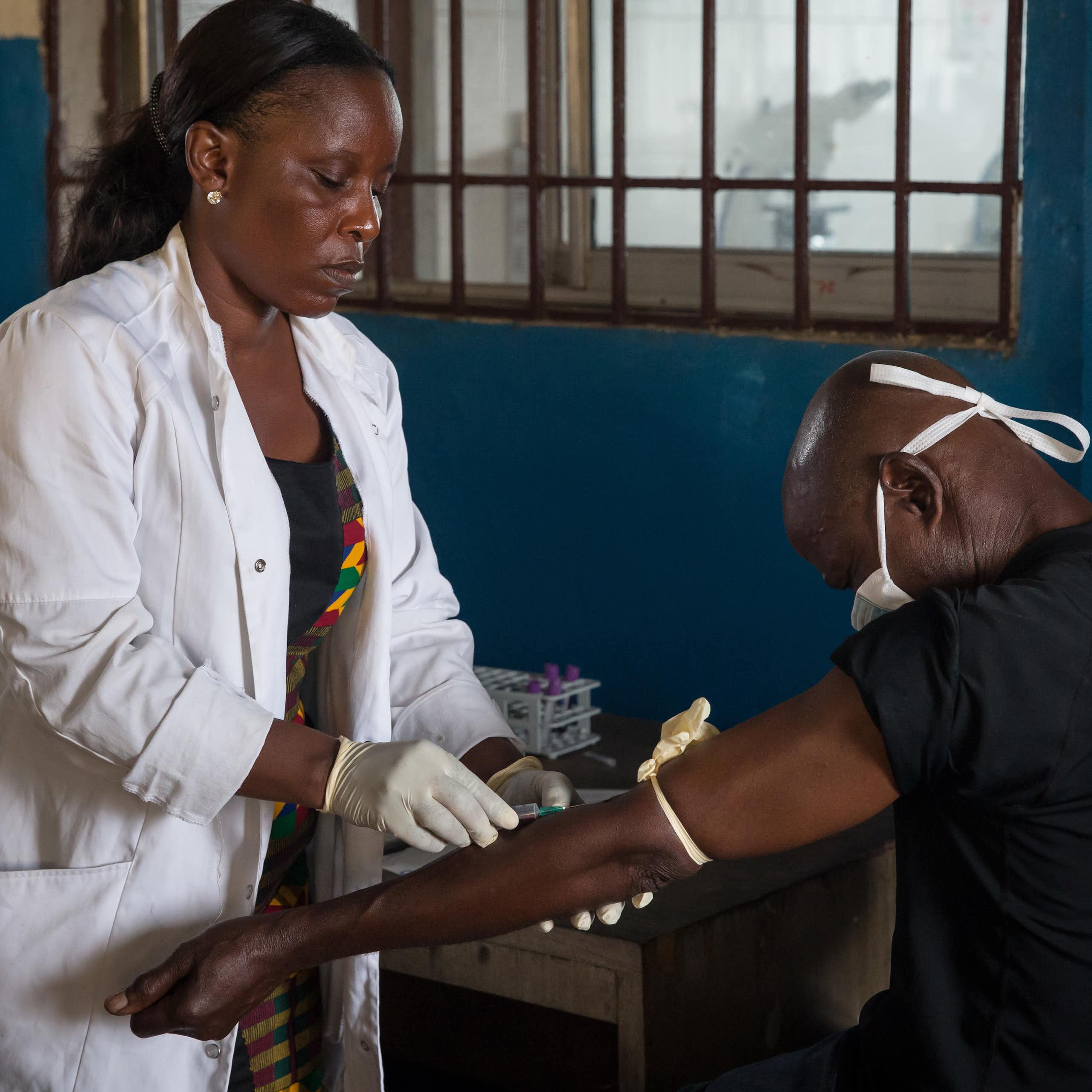 Woman in a white coat and gloves holds the arm of a man who is wearing a face mask