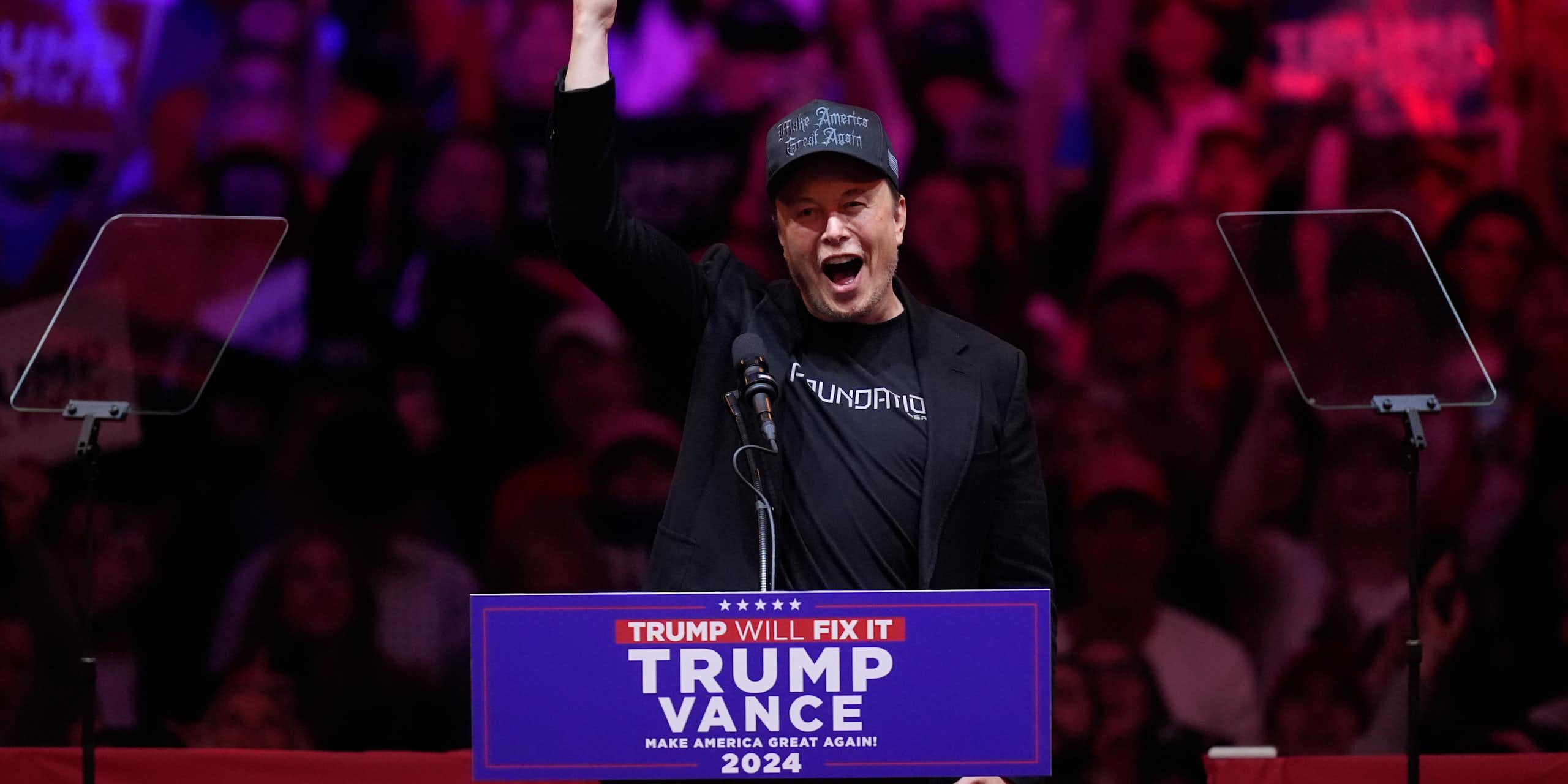 Man wearing black Make America Great Again hat and a black jacket, with his hand raised and mouth open at a lectern.