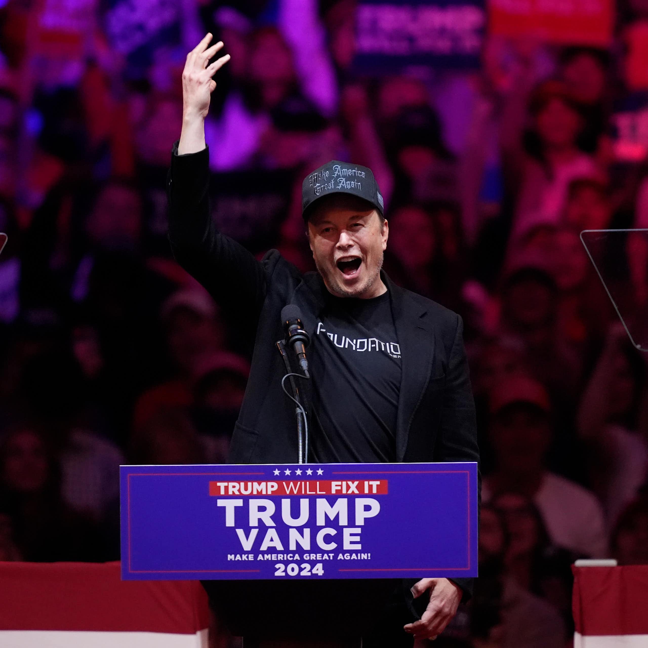 Man wearing black Make America Great Again hat and a black jacket, with his hand raised and mouth open at a lectern.