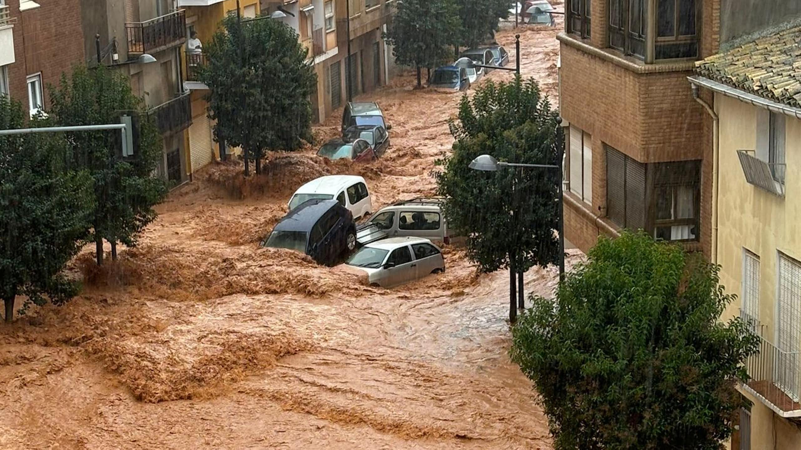 A photo taken from a high window of brown flood waters flowing through a town street, with cars being swept along by the current. 