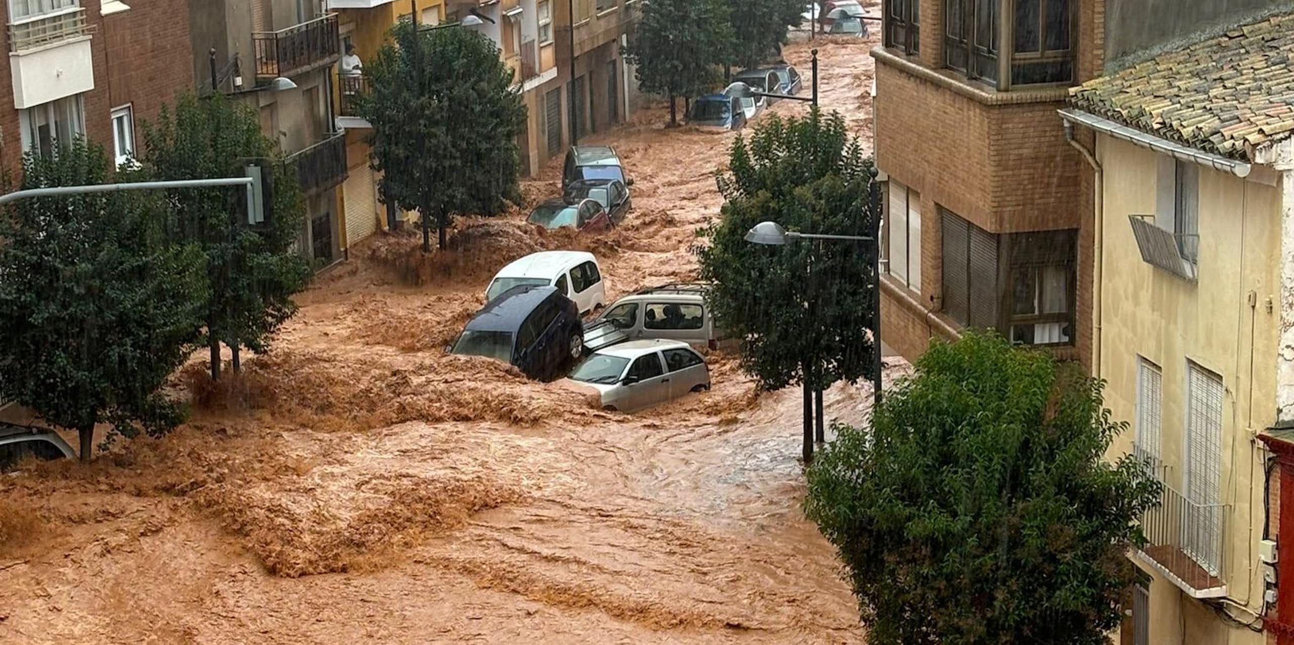 A photo taken from a high window of brown flood waters flowing through a town street, with cars being swept along by the current. 