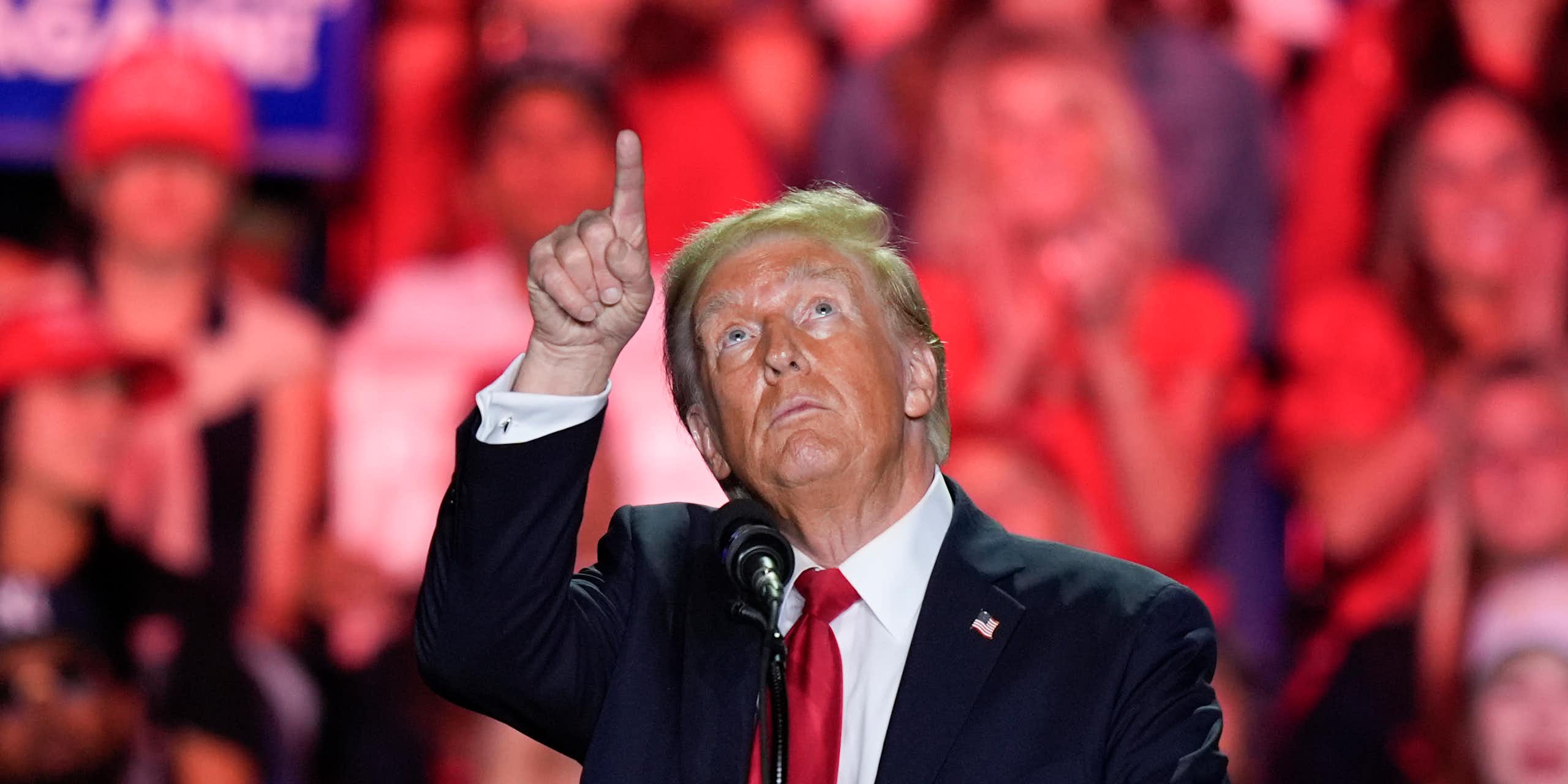 A man with unevenly applied face bronzer and greyish blond hair points up at the ceiling as supporters stand behind him.