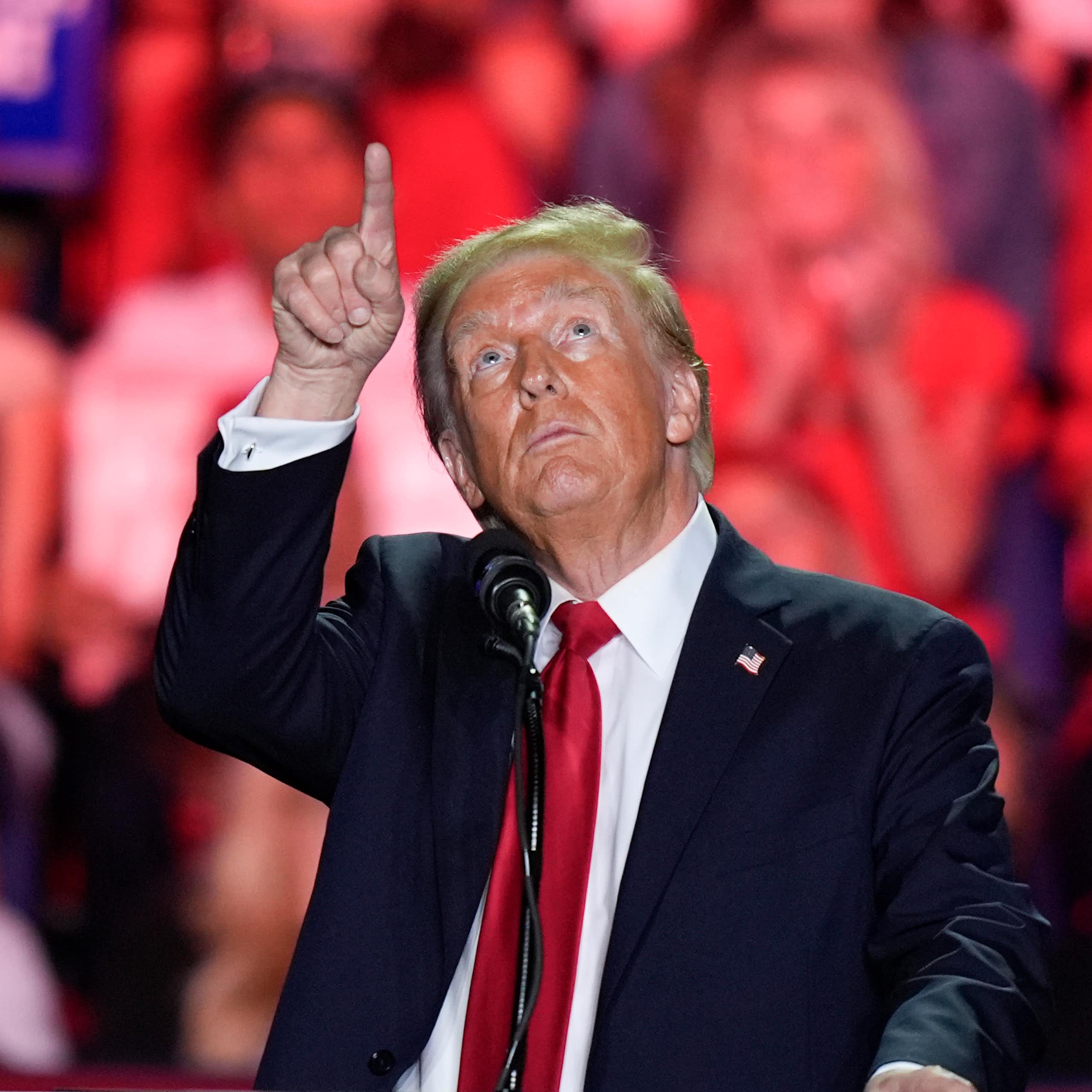 A man with unevenly applied face bronzer and greyish blond hair points up at the ceiling as supporters stand behind him.