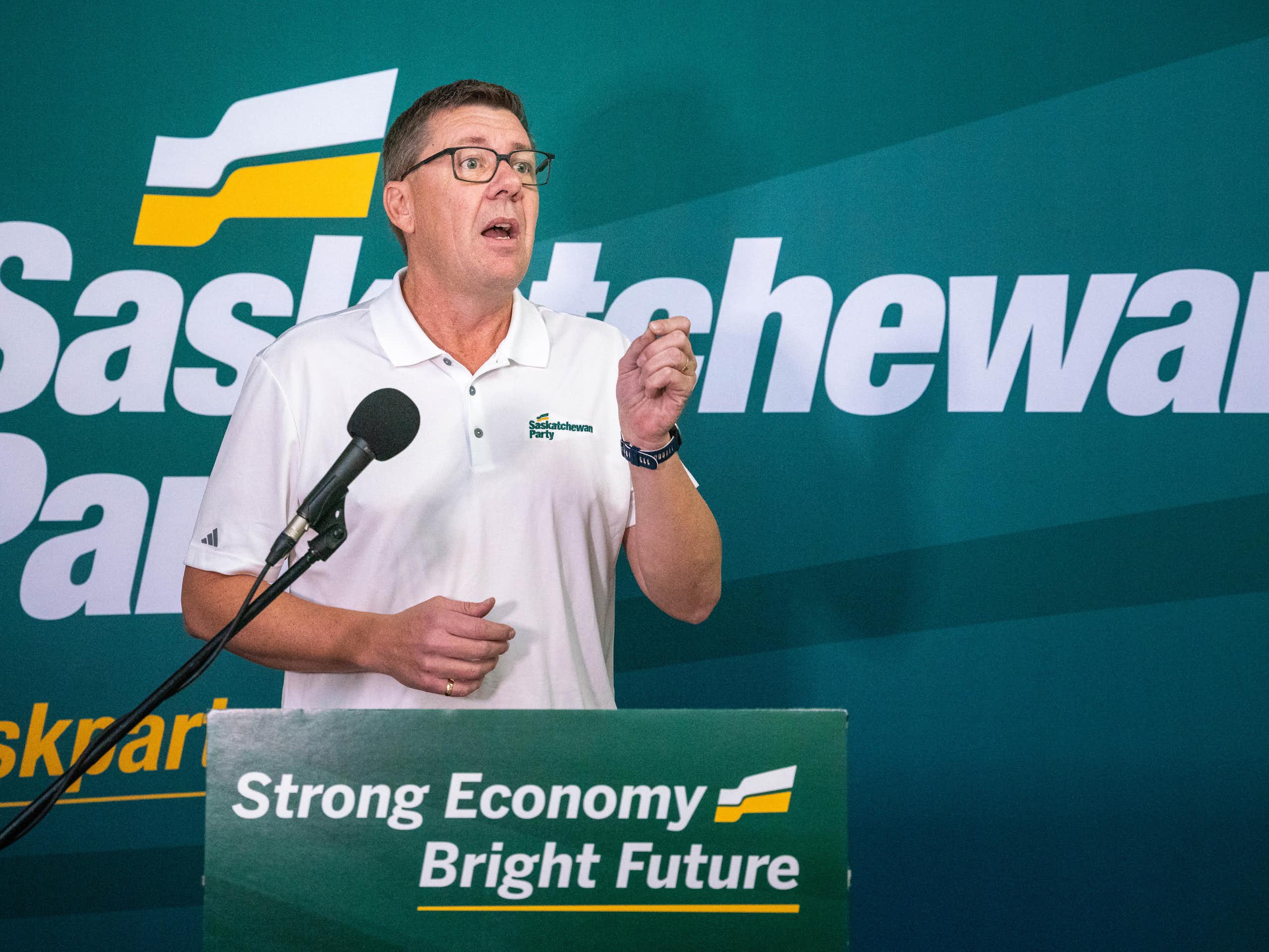 A man in a white collared T-shirt and glasses speaks into a mircophone. A green banner behind reads Saskatchewan Party; the banner on the podium reads Strong Economy, Bright Future.