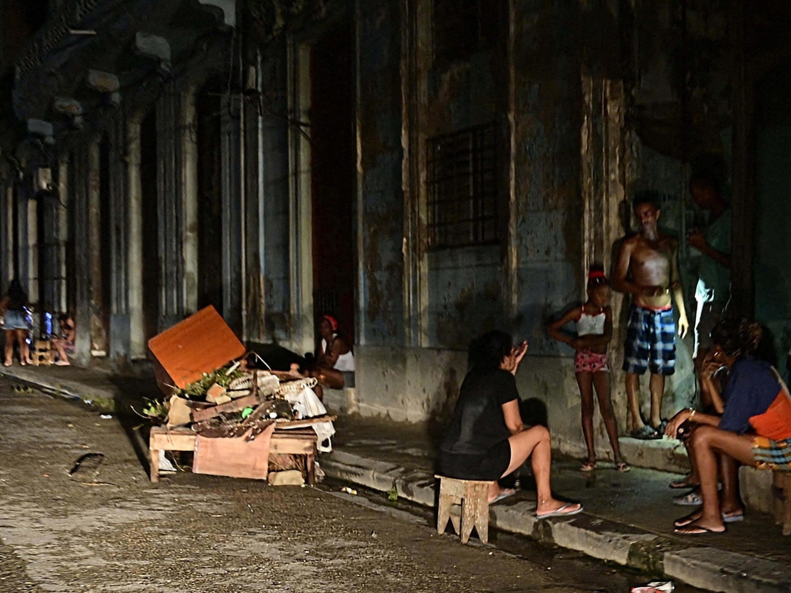 Men and women sit and stand on the side of a road, they are darkly lit.