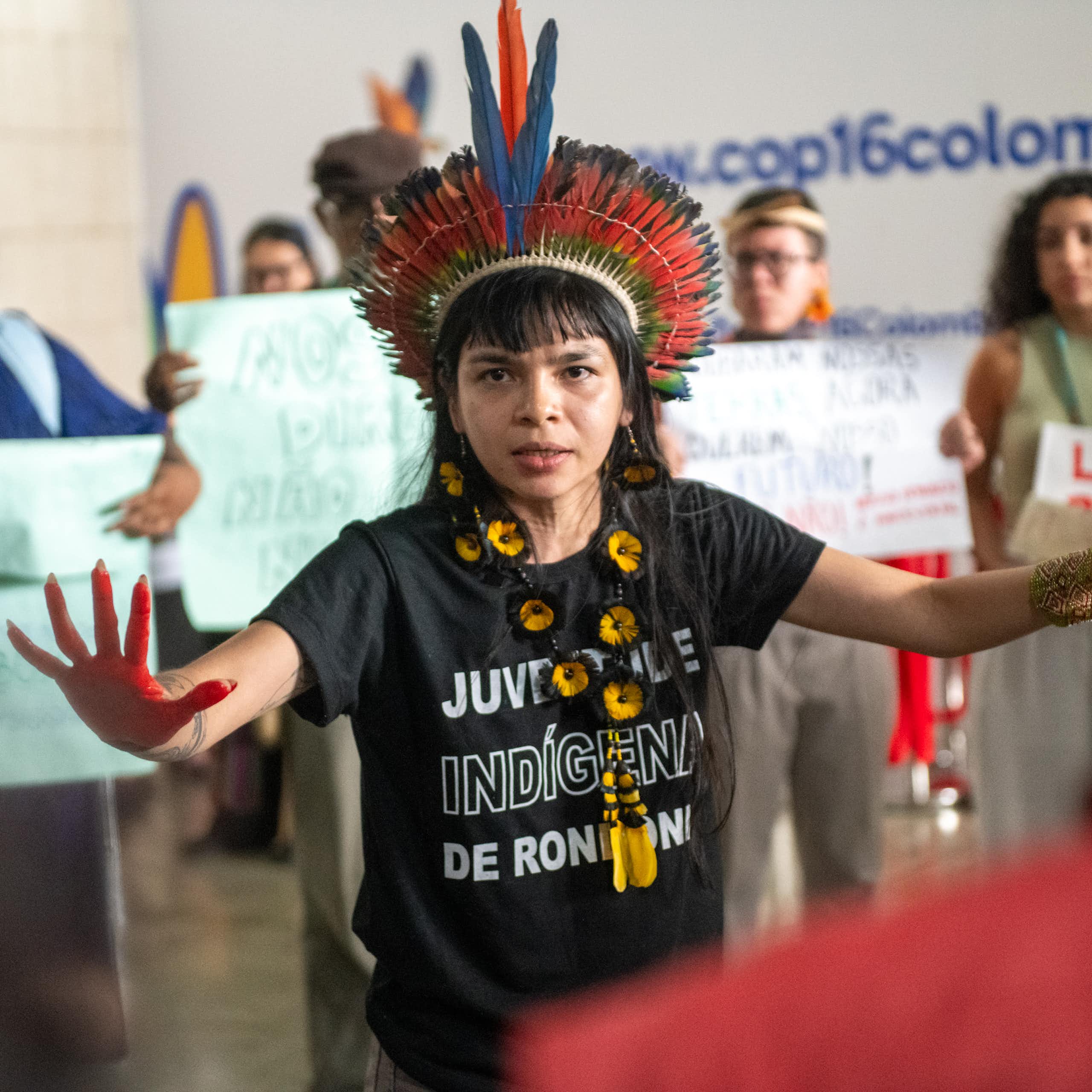woman wearing black T shirt and colourful feathered traditional head dress, hands wide out, women holding signs in background