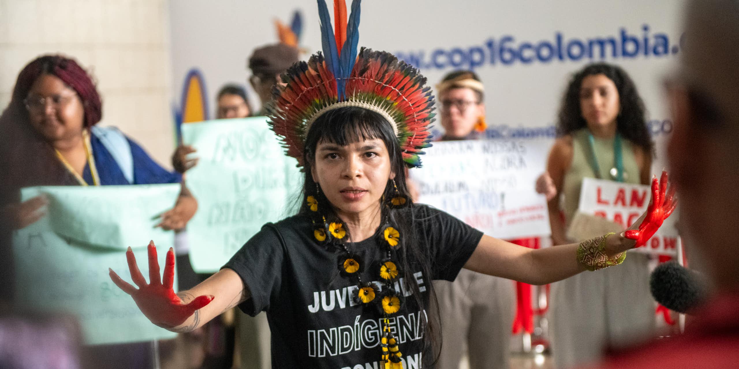 woman wearing black T shirt and colourful feathered traditional head dress, hands wide out, women holding signs in background