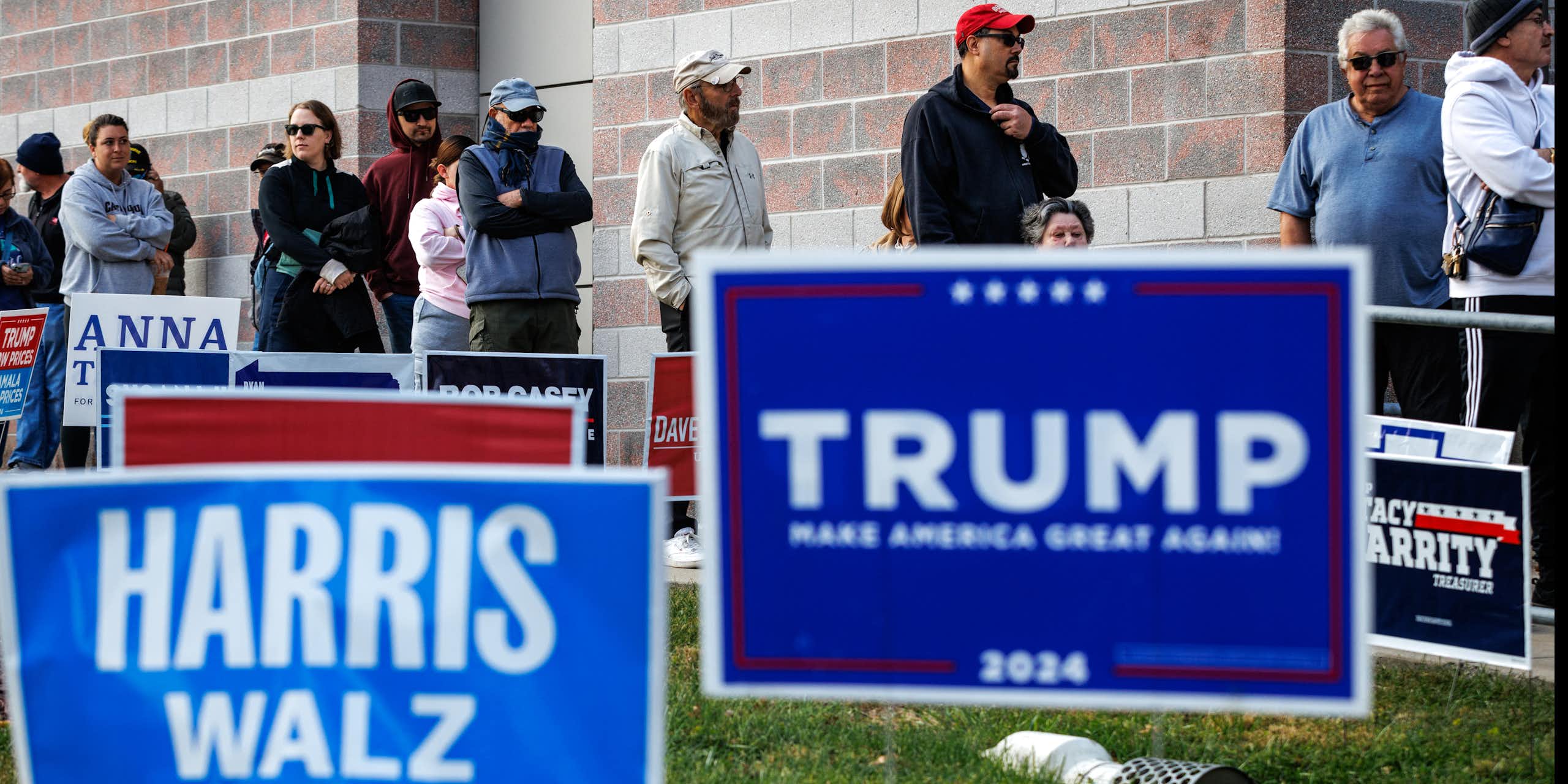 People stand in a line against a building. In front of them are large lawn signs, including two blue ones that say 'Harris Walz' and 'Trump.'