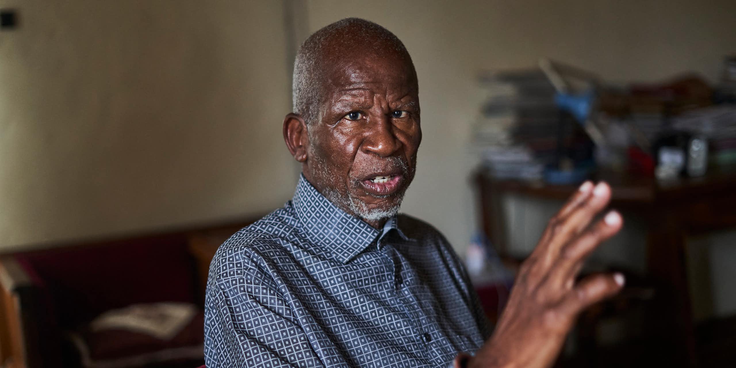 An elder African man with a grey beard wears a blue collared shirt and gestures wirth one hand as he talks.