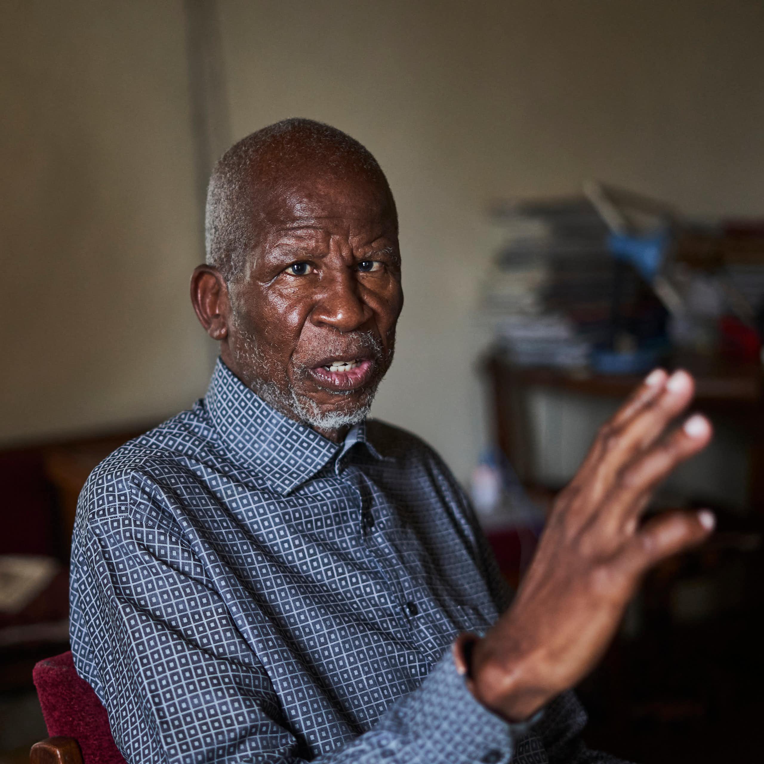 An elder African man with a grey beard wears a blue collared shirt and gestures wirth one hand as he talks.