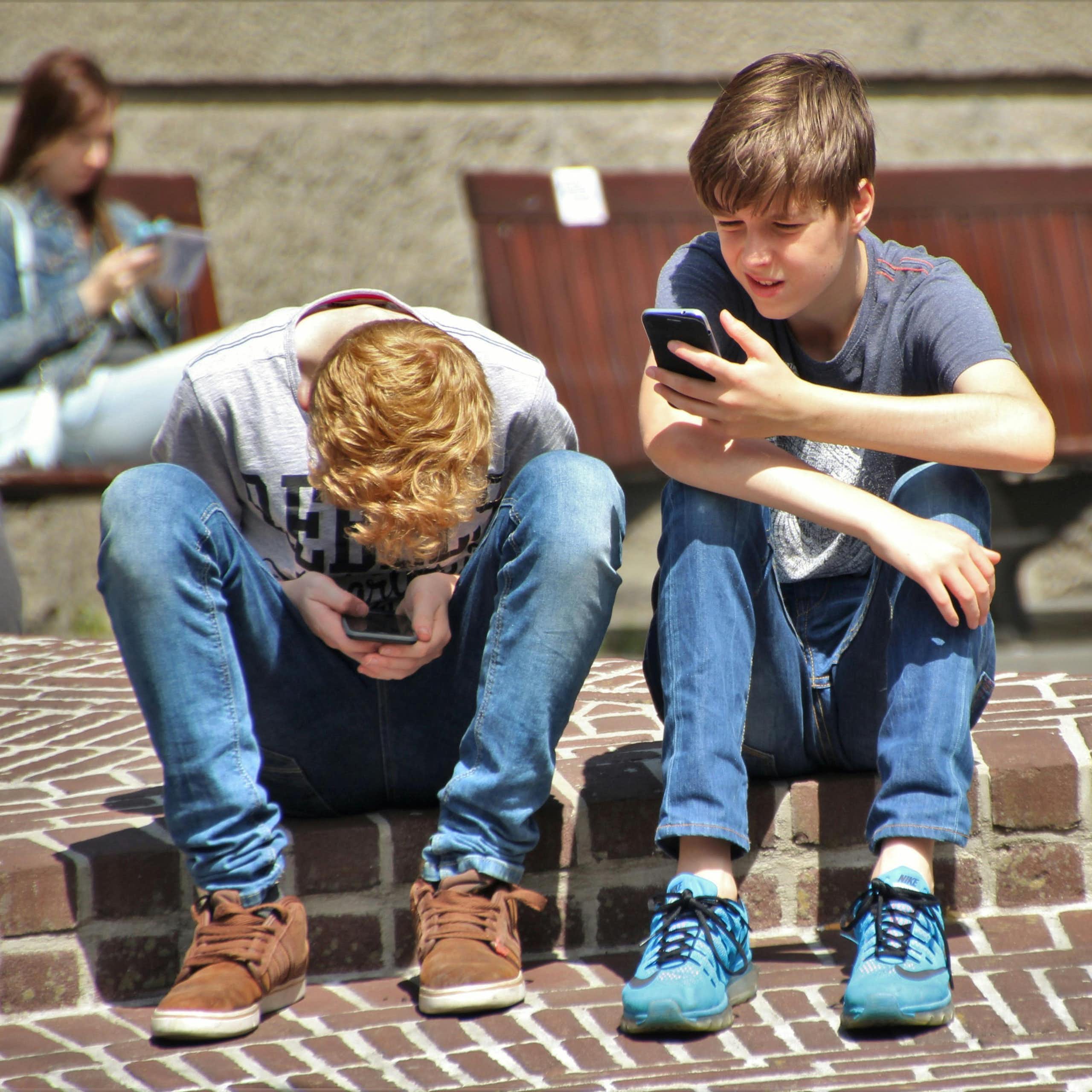 Two young boys in jeans sit on a kerb outdoors looking at their phones.