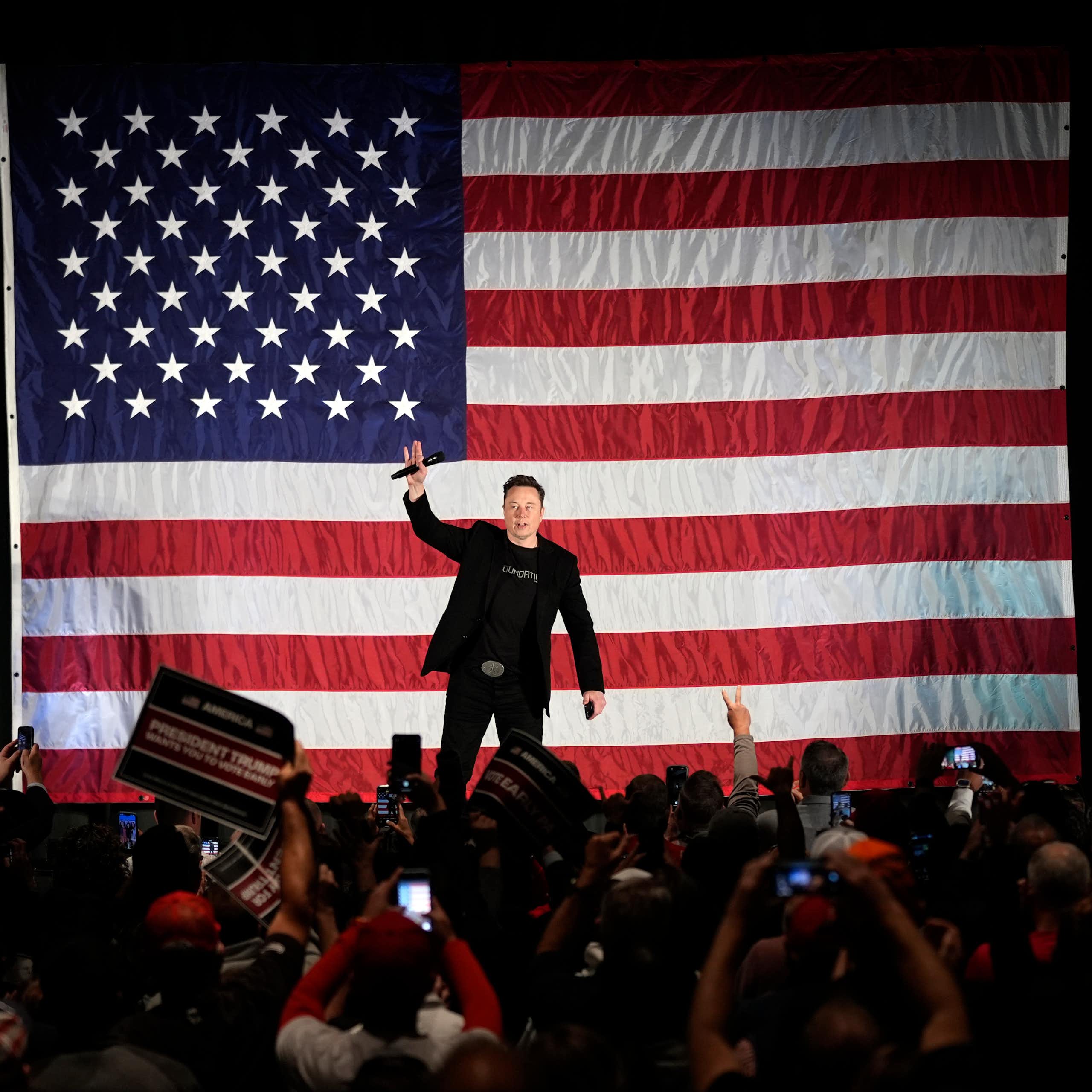 Crowd of people taking photos of a man on stage holding a microphone in front of a large American flag.